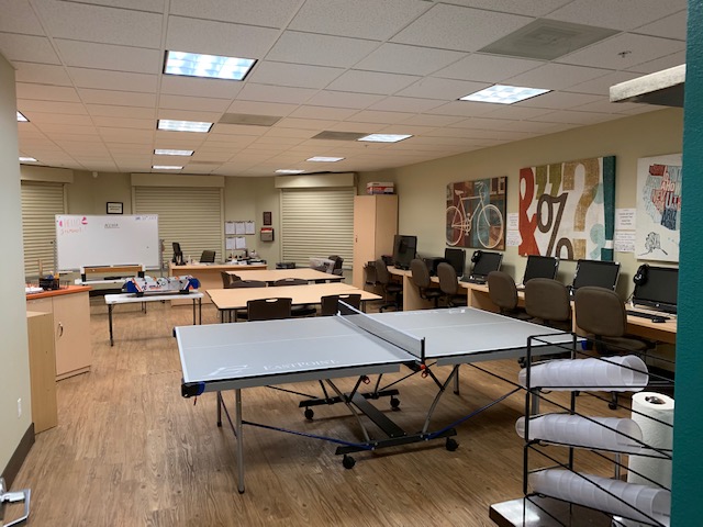 View of family Resource center, multiple tables, racks with supplies, computer station on the right side, three windows with horizontal blinds, white board, cabinet shelves, multiple picture frames on the wall.