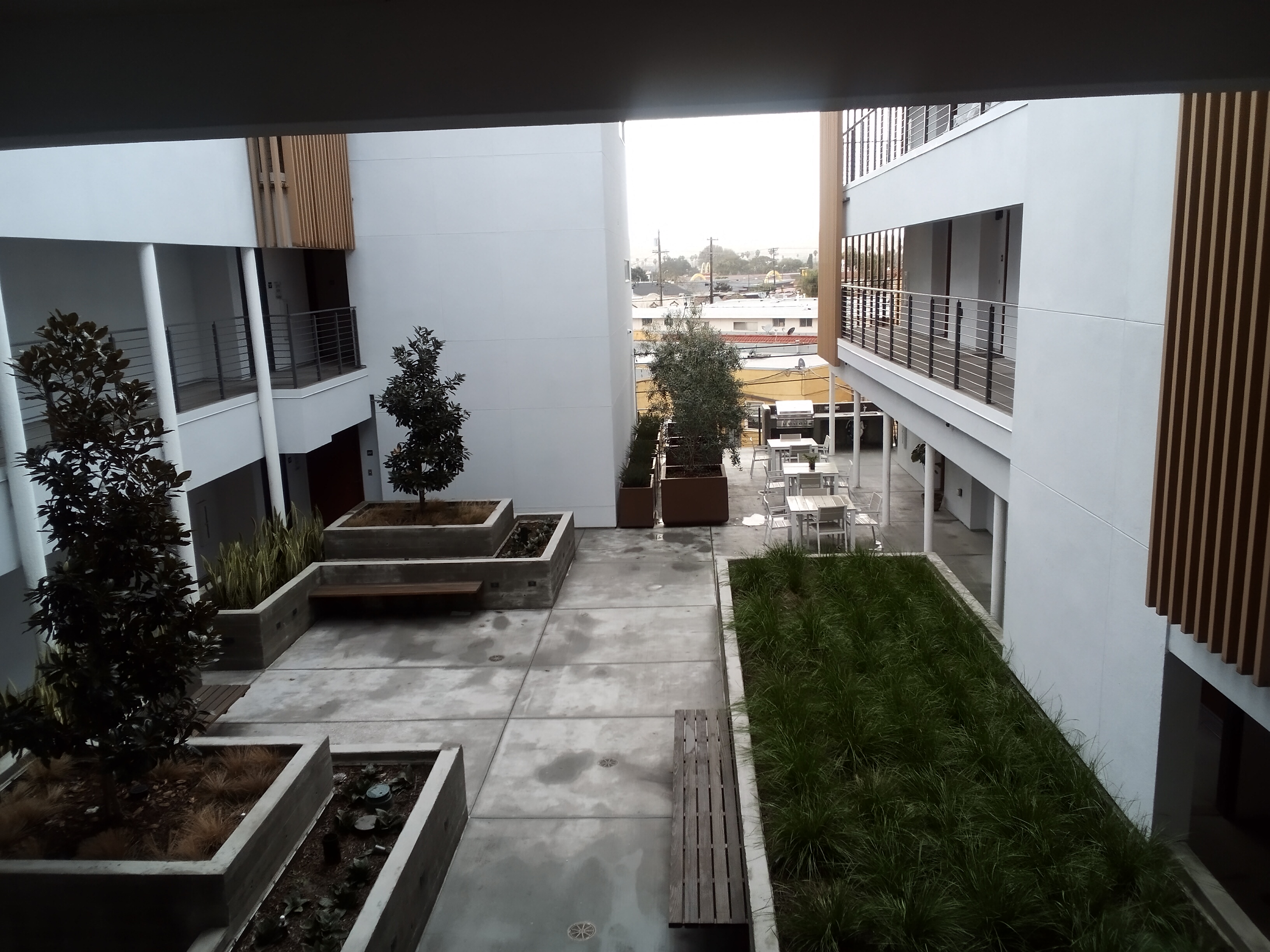 Top View of courtyard. Planters on the perimeter with brown benches. At the far end there are three tables with chairs beside a grill.