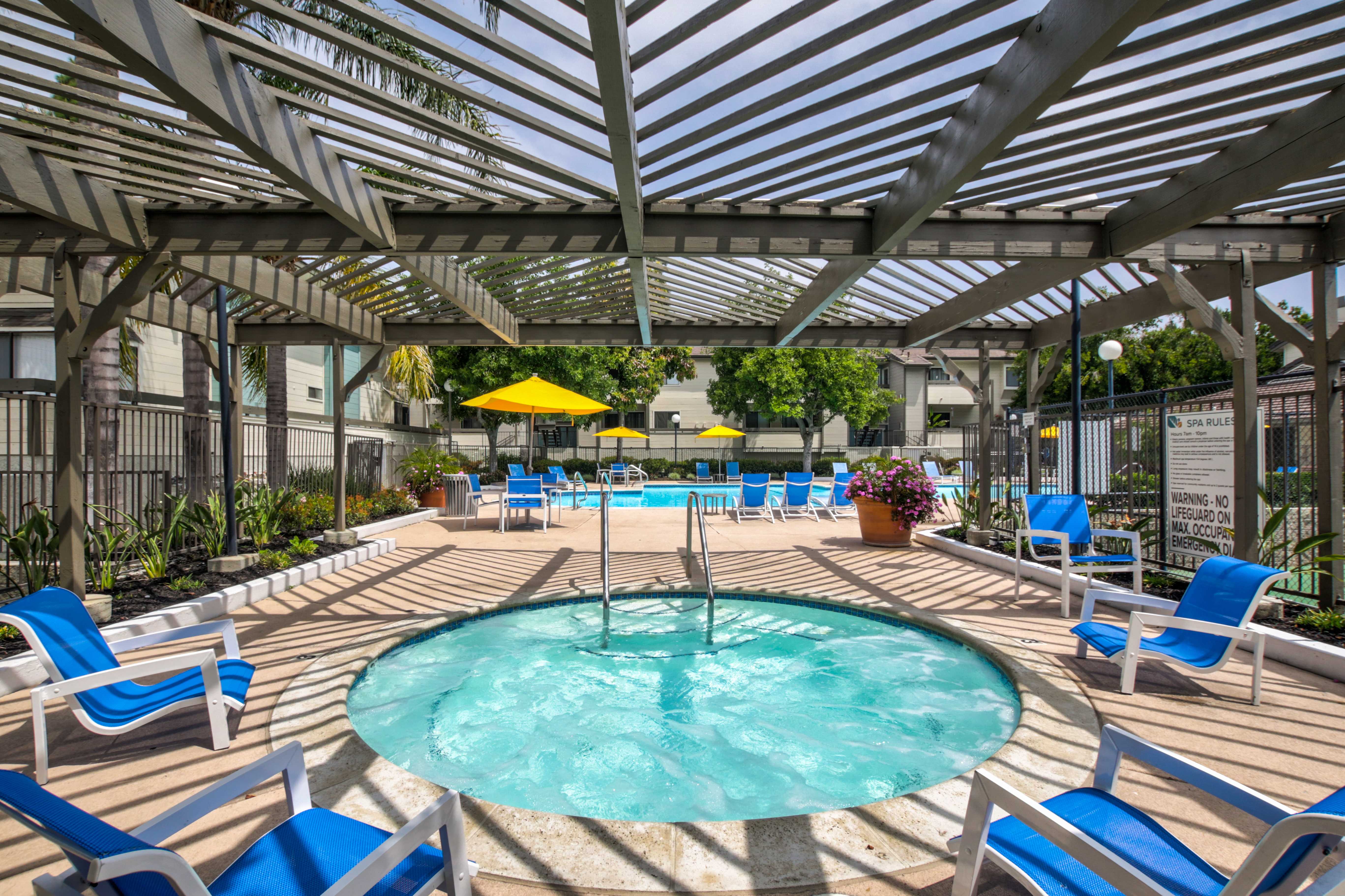 Different view of pool area. Hot tub and a pool a few steps away are visible. There are multiple blue lounging chairs and plants surrounding the area. There are also a few yellow umbrellas for shade.