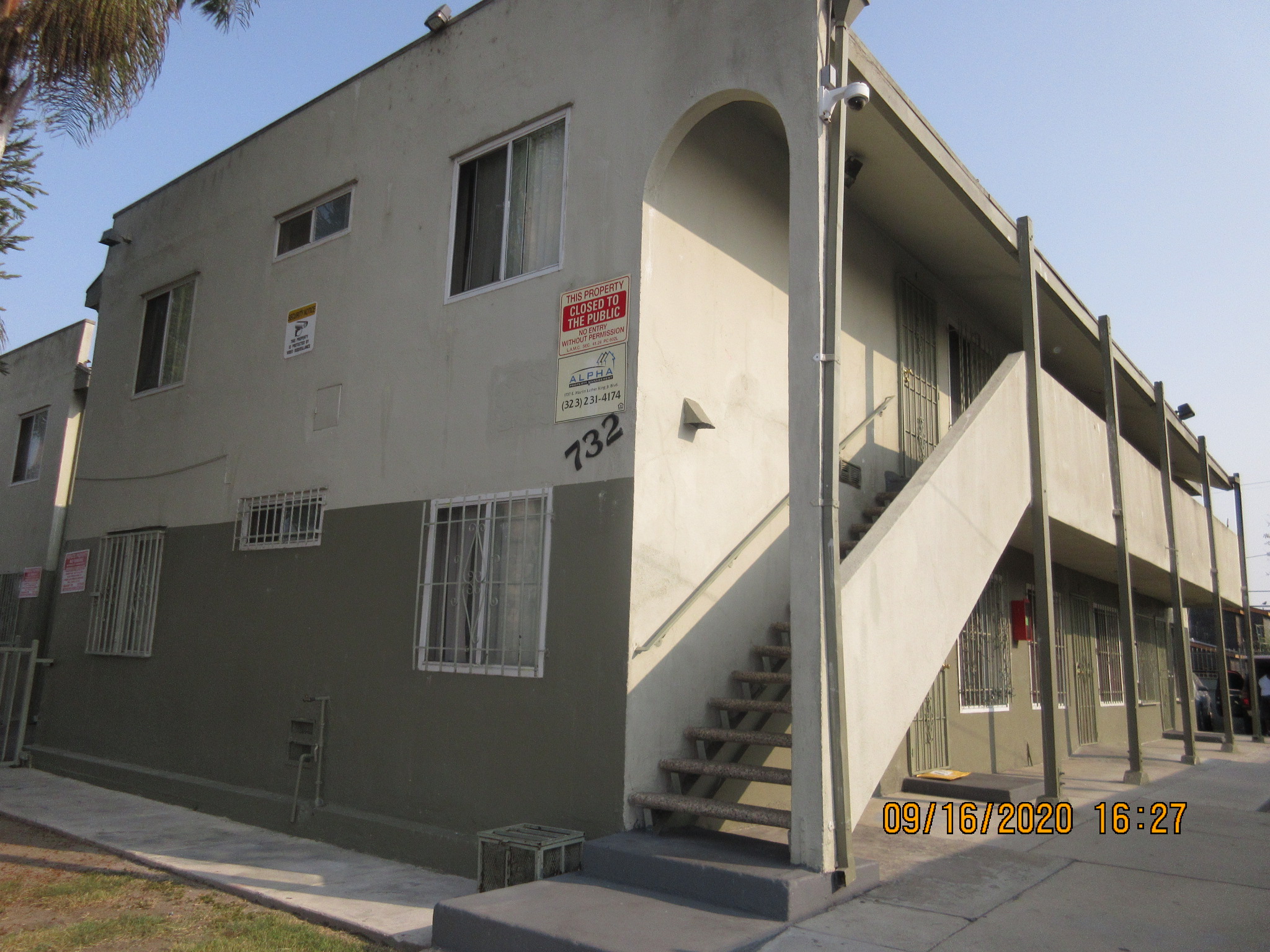 Side view of a a beige and moss green, two story building, multiple windows, gated windows on the first floor, iron screen doors on each unit, stairs up to the second floor one side a wood handrail and the other side a concrete side wall, closed to the pu