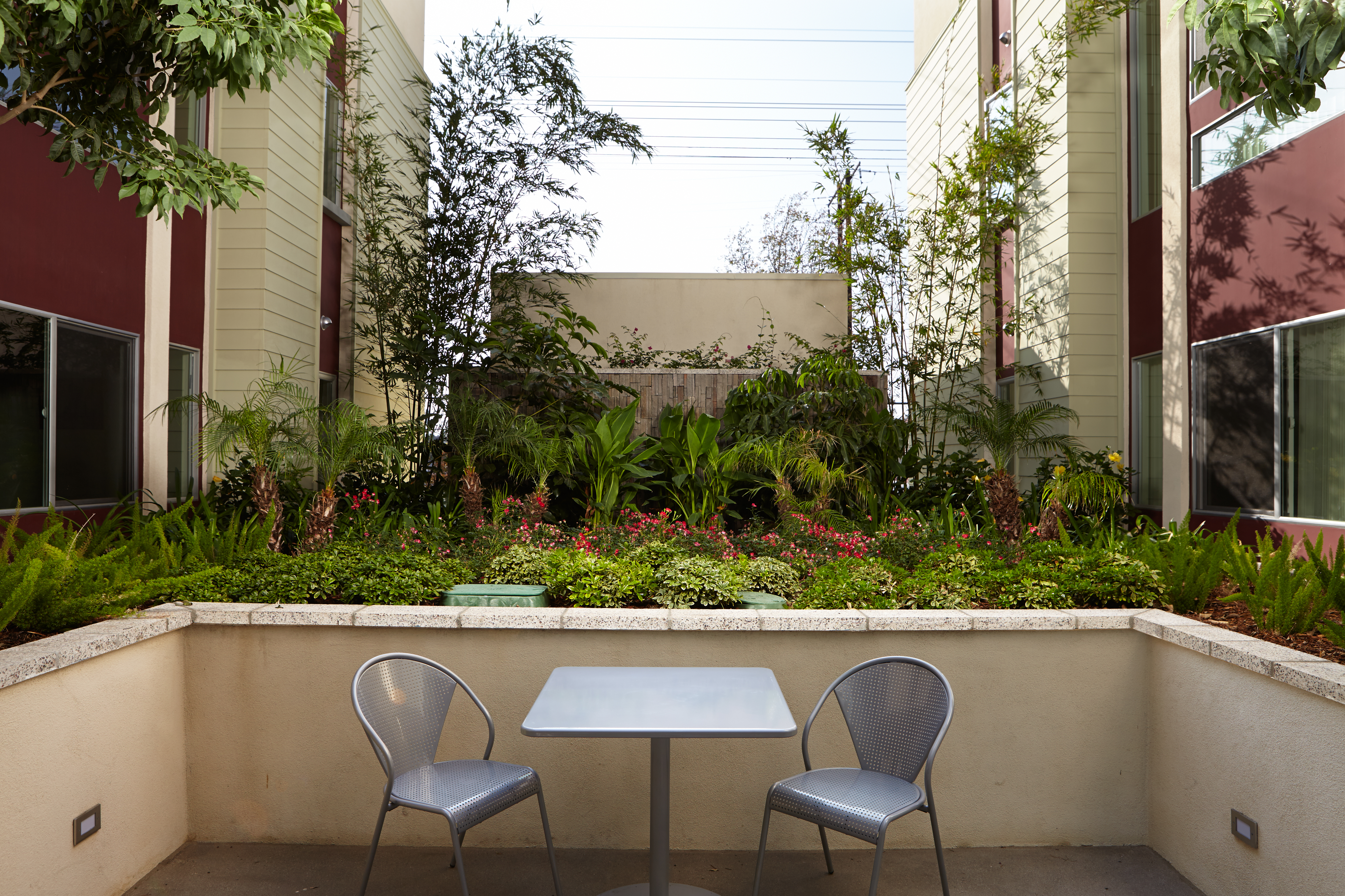 Front view of an outdoor seating area, small square table with two chairs, plants and flower beds behind.