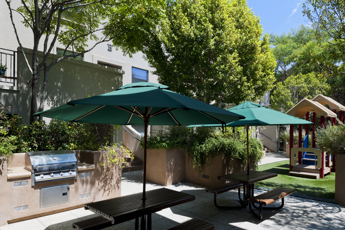 View of a patio area with bench tables, umbrellas, and a grill. Adjacent to it is a small playground, and area is srrounded by plants and trees.