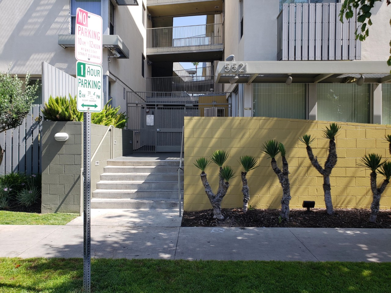 Image of front entrance to Harold Way Apartments. Steps with hand rail leading up to gate entrance of building. On either side of side walk area plant beds and grass. Street parking notification sign near street that states no parking from 10am to 12pm on