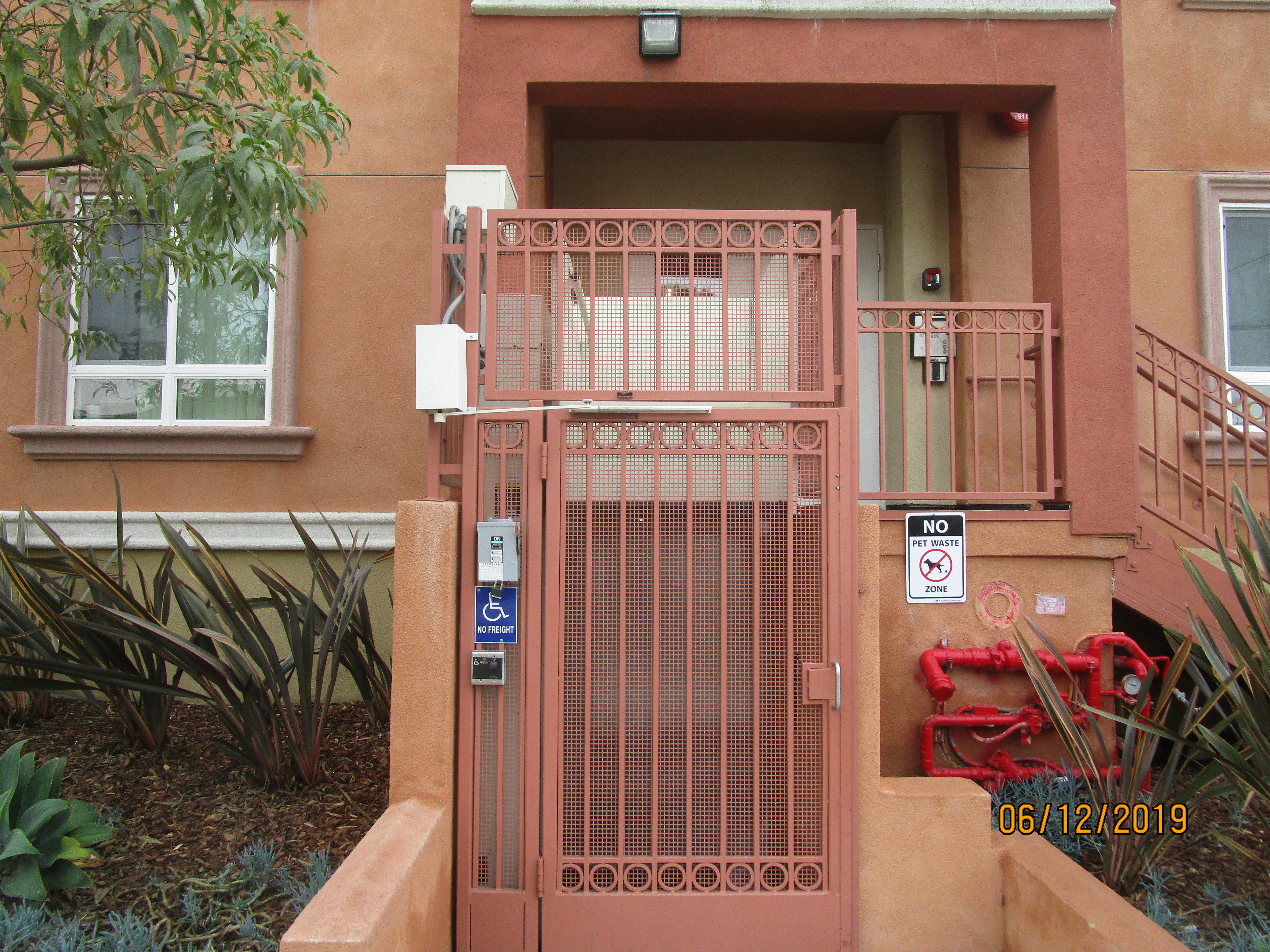 Full front view of a brick color metal locked door, call box and a No Handicap Freight sign on the left side of the door, No pet waste sign on the right side of the door, planters on both side, red water meter on the right.