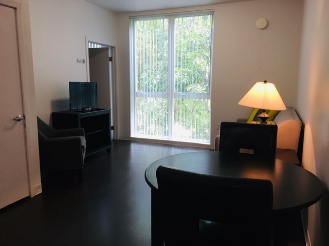 View of a unit, dark brown wood floors, small black round table with two chairs, other couches, a TV, a window with white vertical blinds.