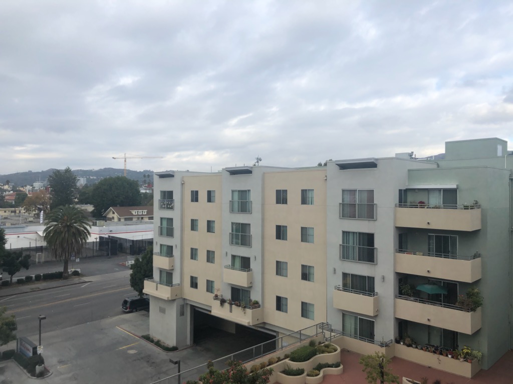 Rooftop view of a four story white and beige building. There is an extra bottom floor designated for parking entrance. Units have balconies, and there is an open space on the second floor that has a variety of plants,