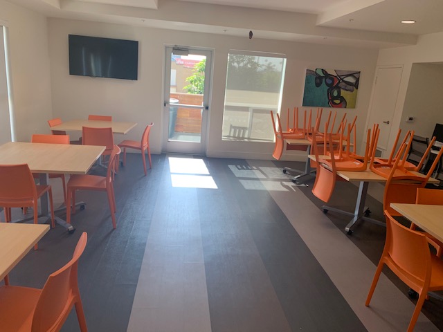 Interior view of a community room that contains multiple tables and chairs. Tables contain wheels under them, but orange chairs do not. There is a flat screen on one of the walls and a painting in another. There is also a computer on the side of the room.