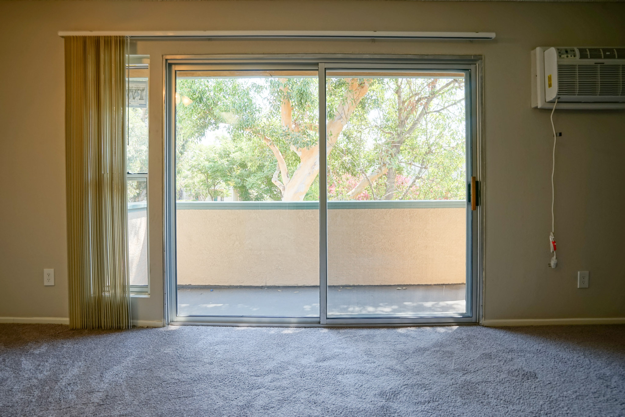 View of a glass sliding door that leads to a balcony. Door has vertical blinds, and there is an air conditioner mounted on the wall next to it. View outside the balcony is a tree from this angle.