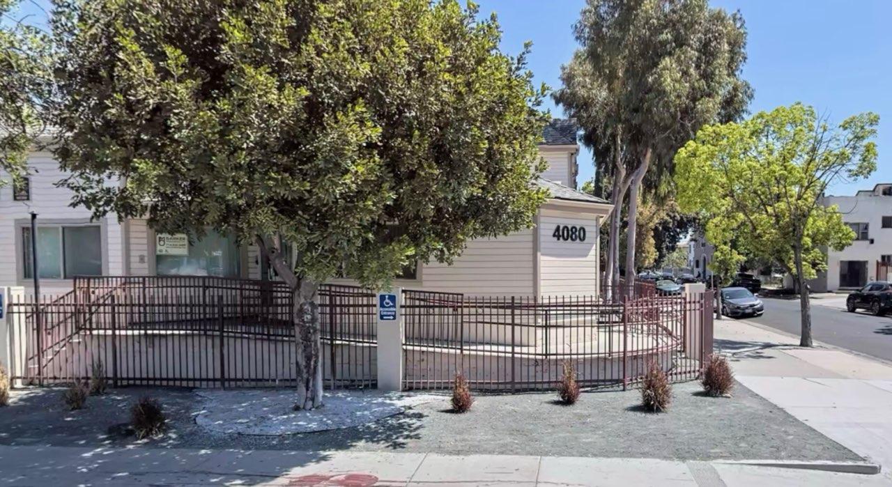 Photo of the apartment, with a fence, on ramp access, trees and parked cars on the street.