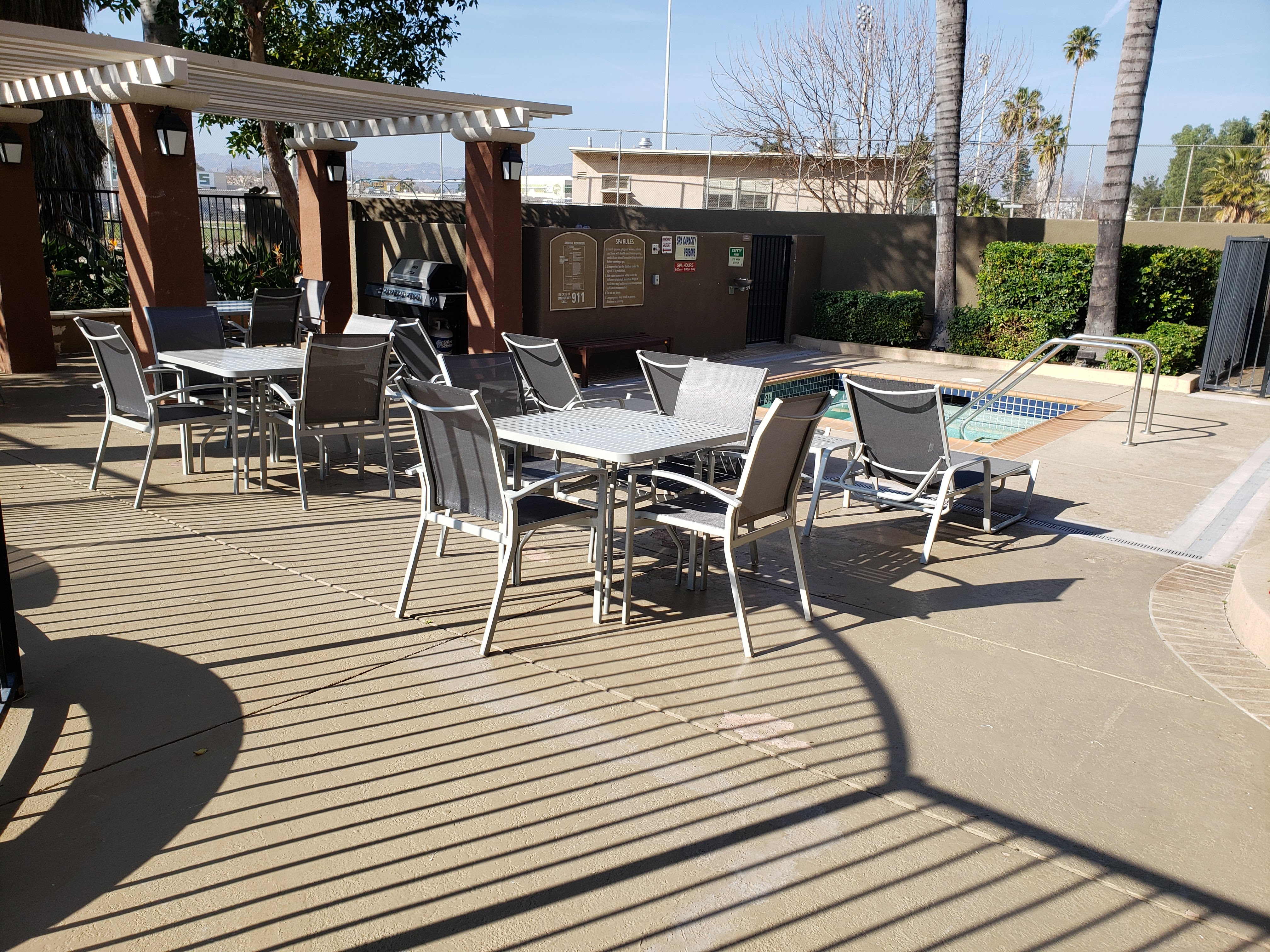 Image of vintage crossing senior apartments outdoor spa area. Are is fenced off. spa located to far right of imgae near bushes and palm trees. Pool shairs next to spa. Behind the pool shairs are patio tables and chairs. bbq grill available under gazebo.