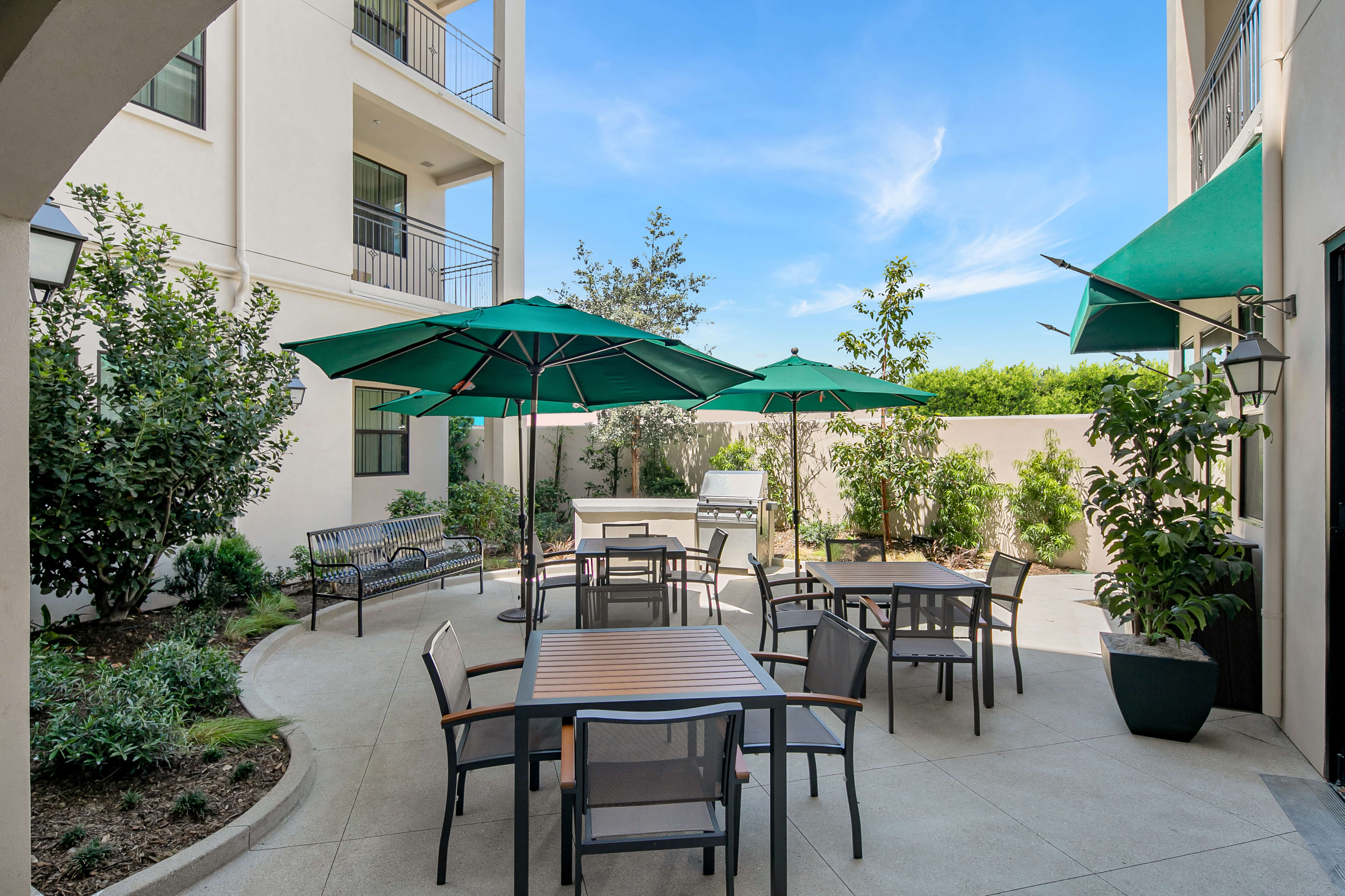 Outdoor patio and barbecue with three tables, two umbrellas, surrounded by flowerbeds.