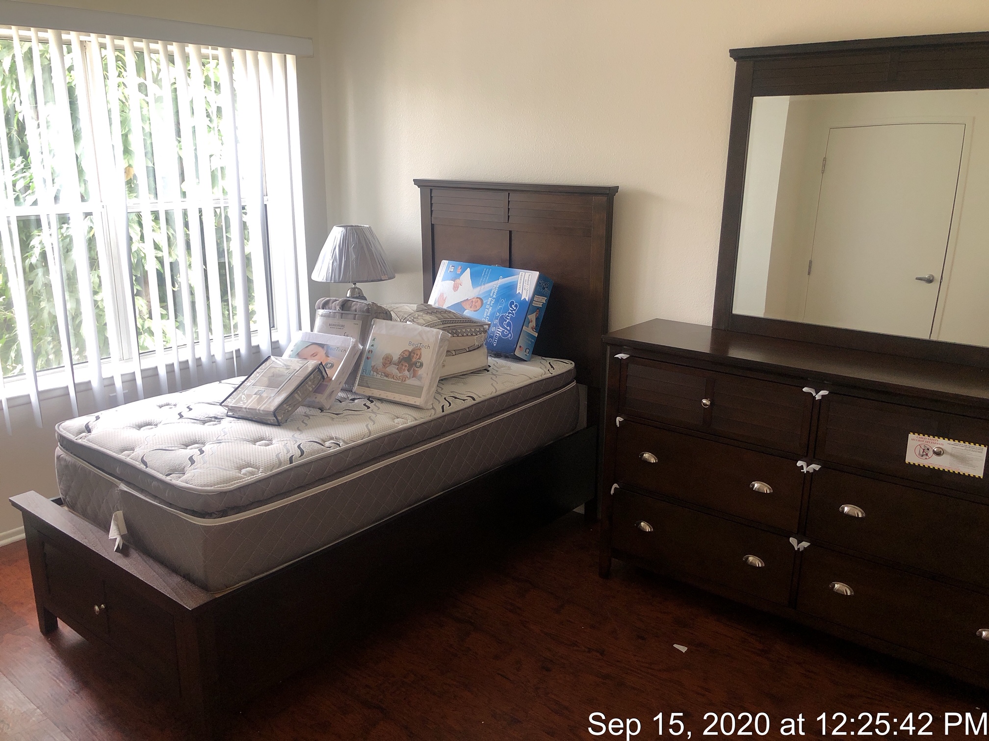 HFL Palms Court - View of a bedroom with wooden floors and a window with blinds. Room has a bed frame with a mattress, a lamp, and a drawer with a mirror.