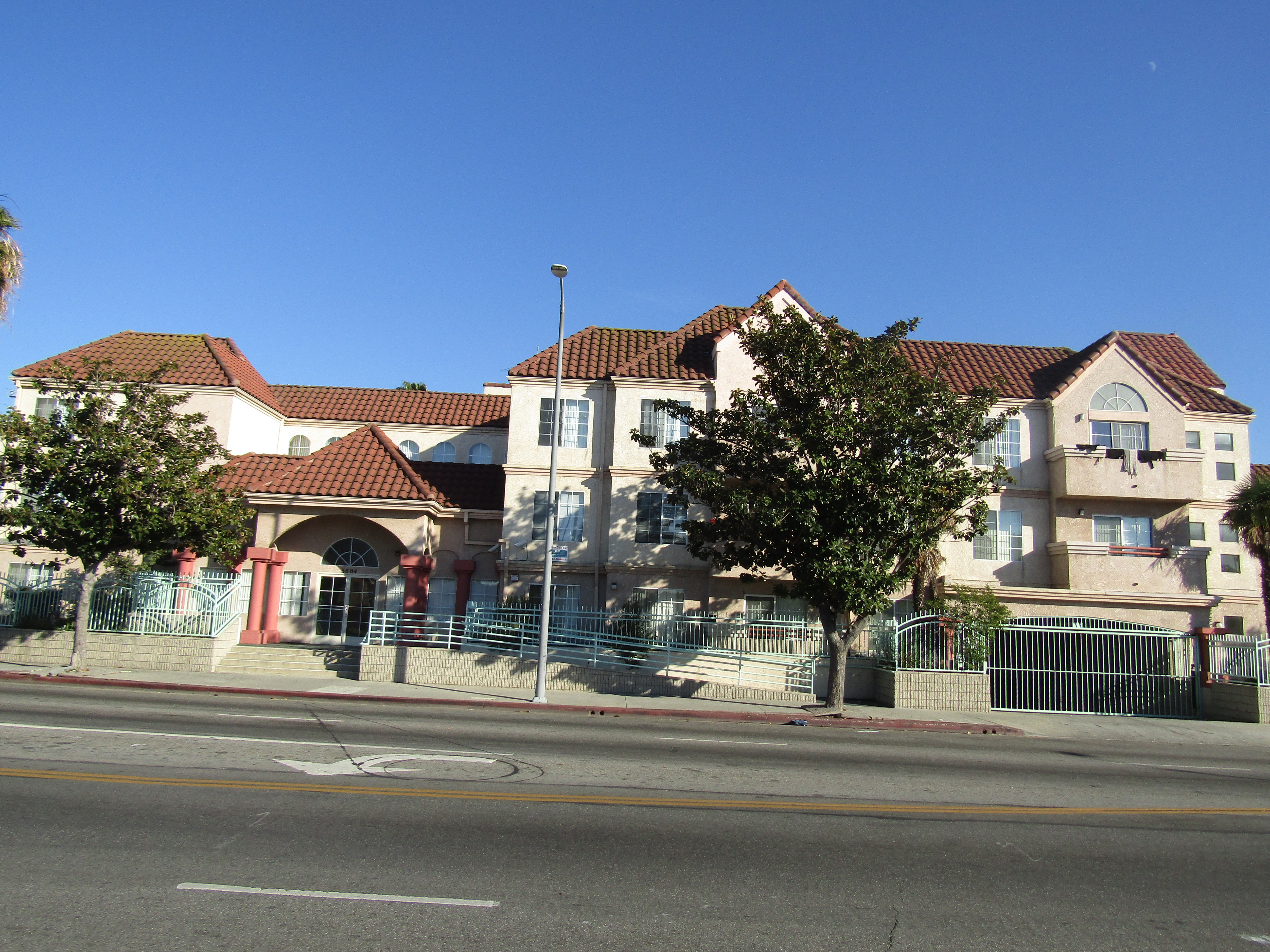 Front view of a three story light brown building. Entrance has stair and ramp access. There is a gated parking entrance. Units facing the street have balconies. There are two trees in front of the building,