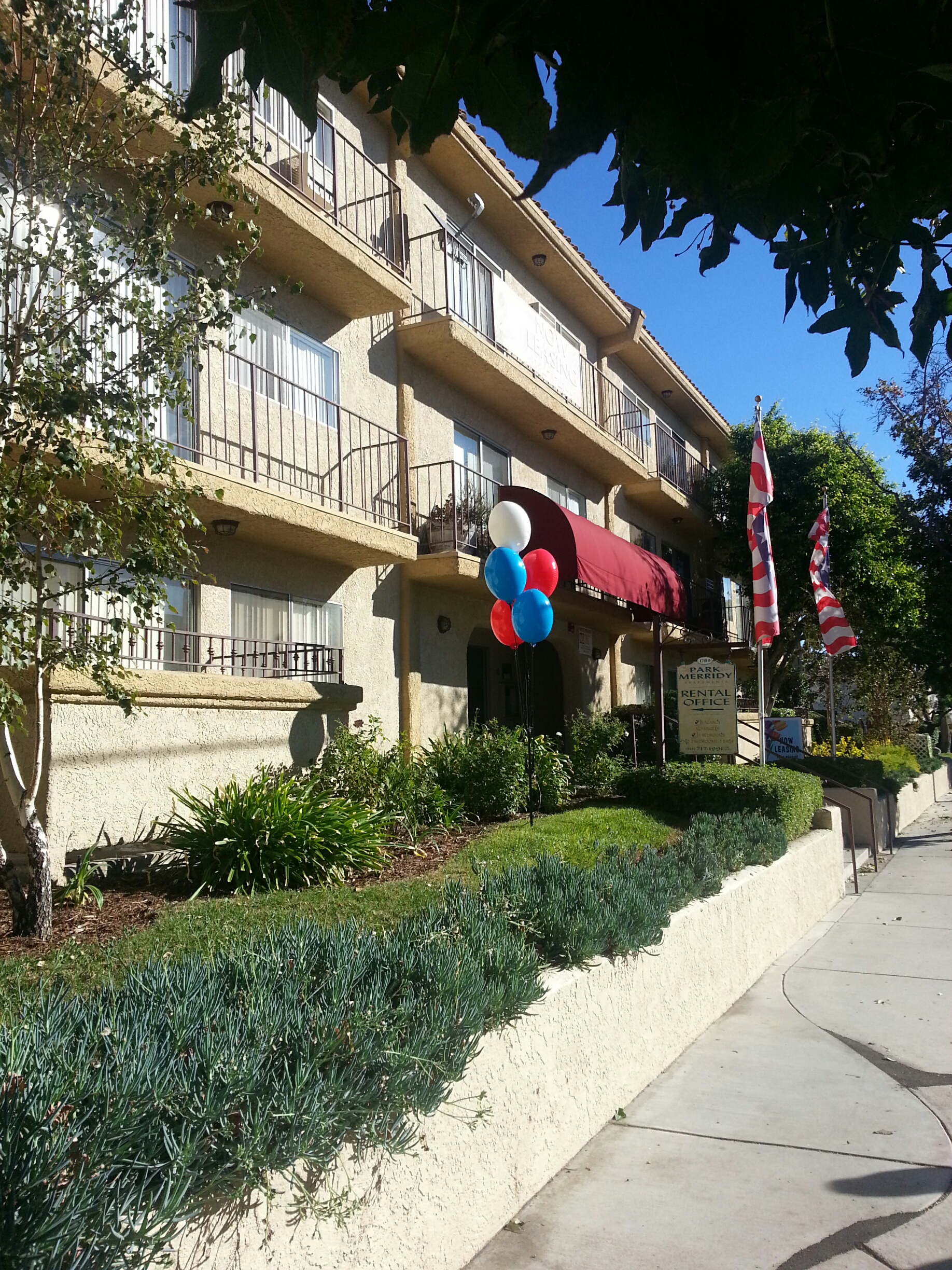 three story light yellow building, burgundy canopy right at the entrance with steps and black steel handrails, blue,white and red ballons by entrance, two USA flags on each side of the handrail, trees and landscaping, Rental and building sign