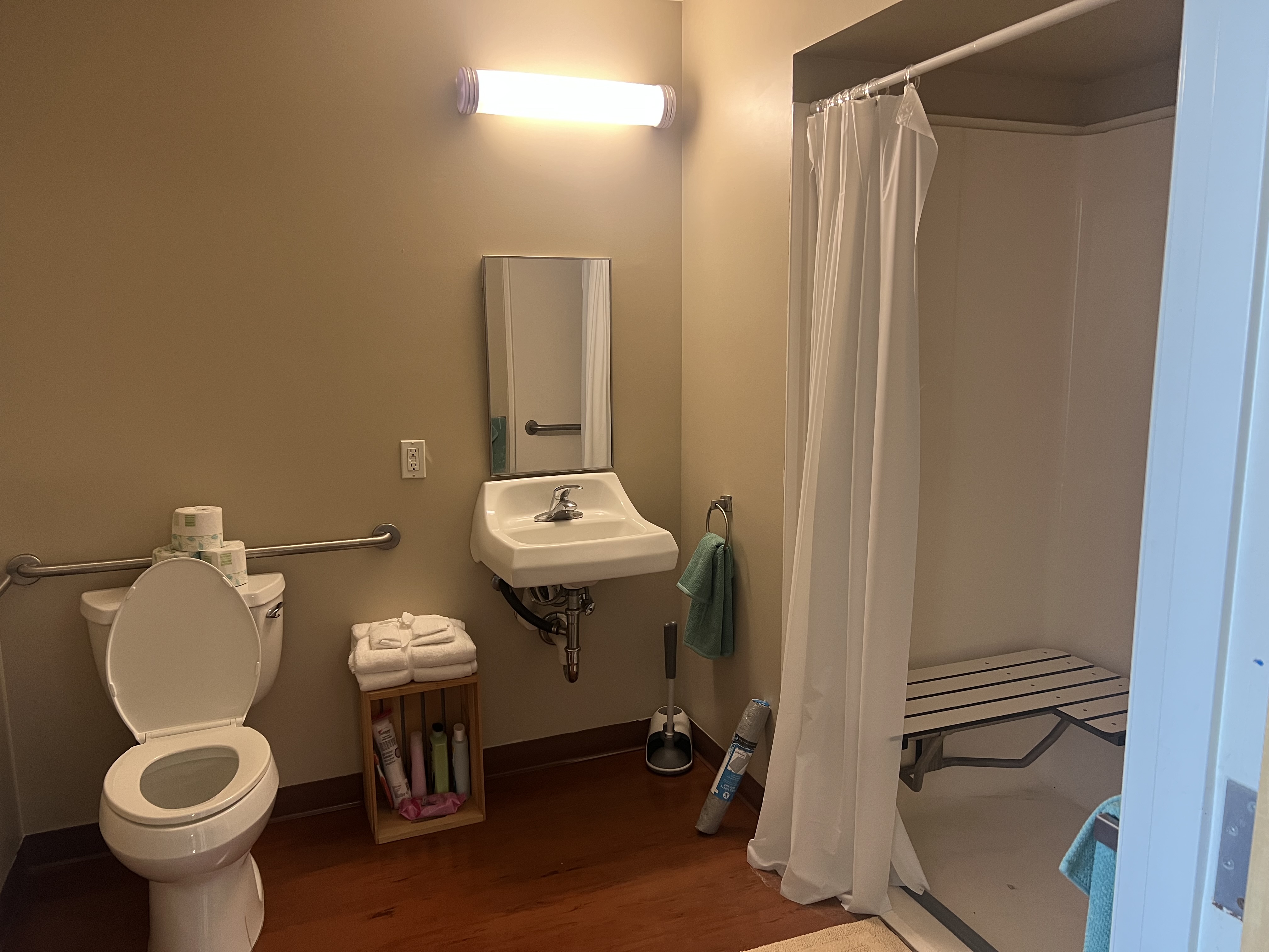 Bathroom with grab bars behind the toilet and roll-in shower with seat. Plenty of linen towels; small mirror over the sink.
