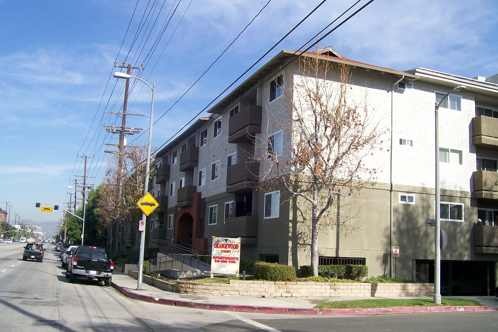 Side view of 3 story whiteand brown apartment building with brown balconies, bushes, trees, street parking along building