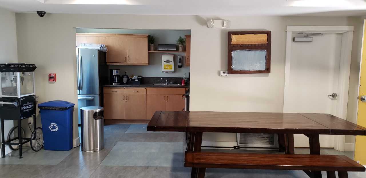 Image of Rayen Apartments community kitchen. Large picnic table with bench seating. In the kitchen there is a large double door fridge. Upper and lower counter tops with large two sided sink. Coffee maker, paper towel dispencer located neat cabinets. Port