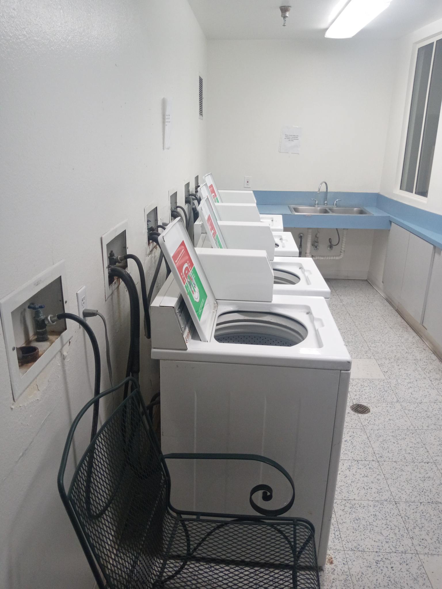 Laundry room. Four washing machines are lined against the wall. There is a sink and a chair.