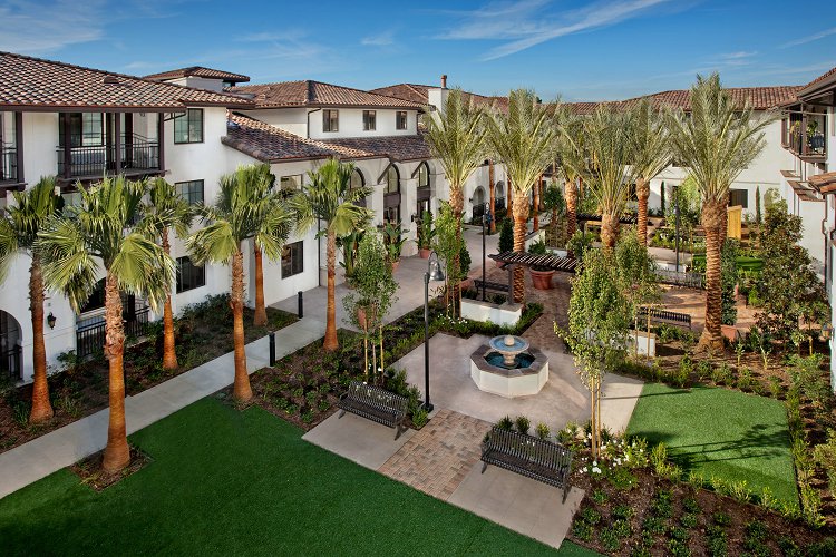Aerial view of a courtyard inside a three story property. Area has multiple palm trees, bushes and plants. There's a water fountain located in the middle, and benches around it. There is also a large grass area.