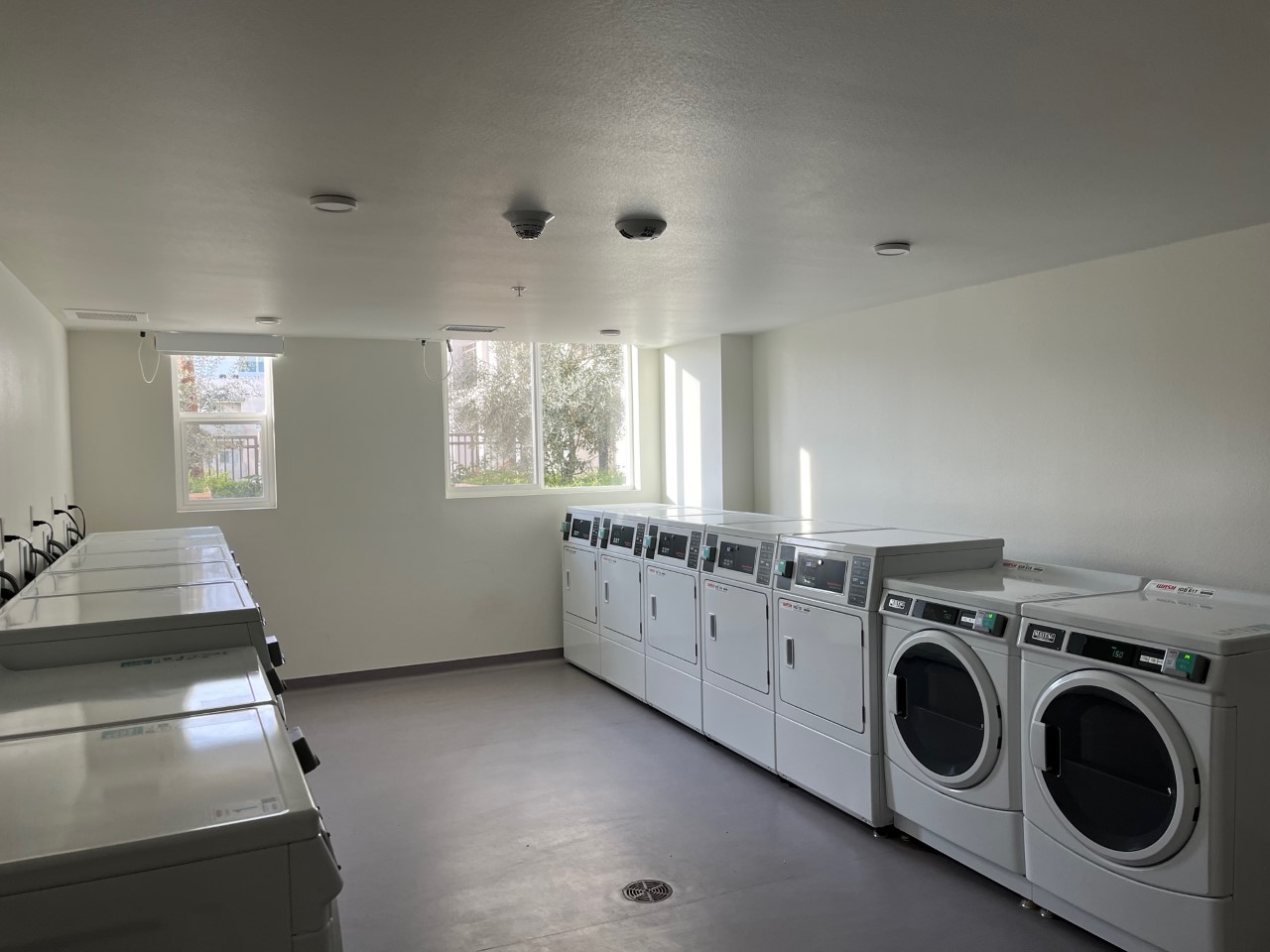 Laundry room with two rows of machines facing each other. On the far wall there are two windows with a view of the courtyard.
