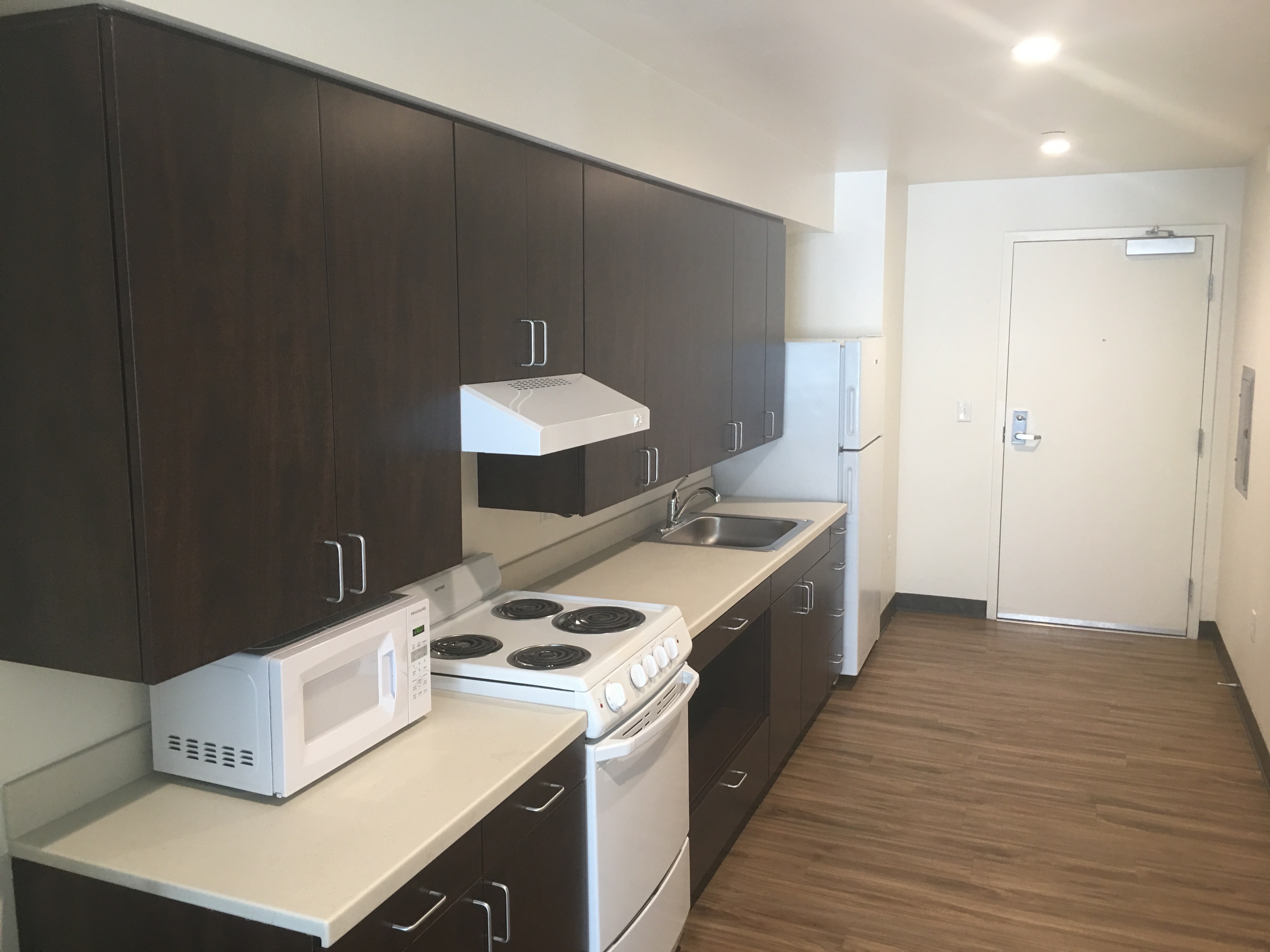 Interior view of a kitchen in a unit at the PSH Campus. Dark brown cabinets with white countertops, microwave, stove and refrigerator.