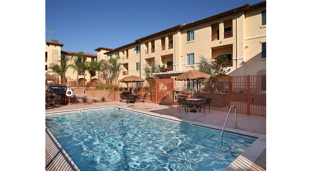 View of an outdoor pool with two table sets with umbrellas. Area is fenced around. Building is a three story complex, and some units have balconies facing the pool.