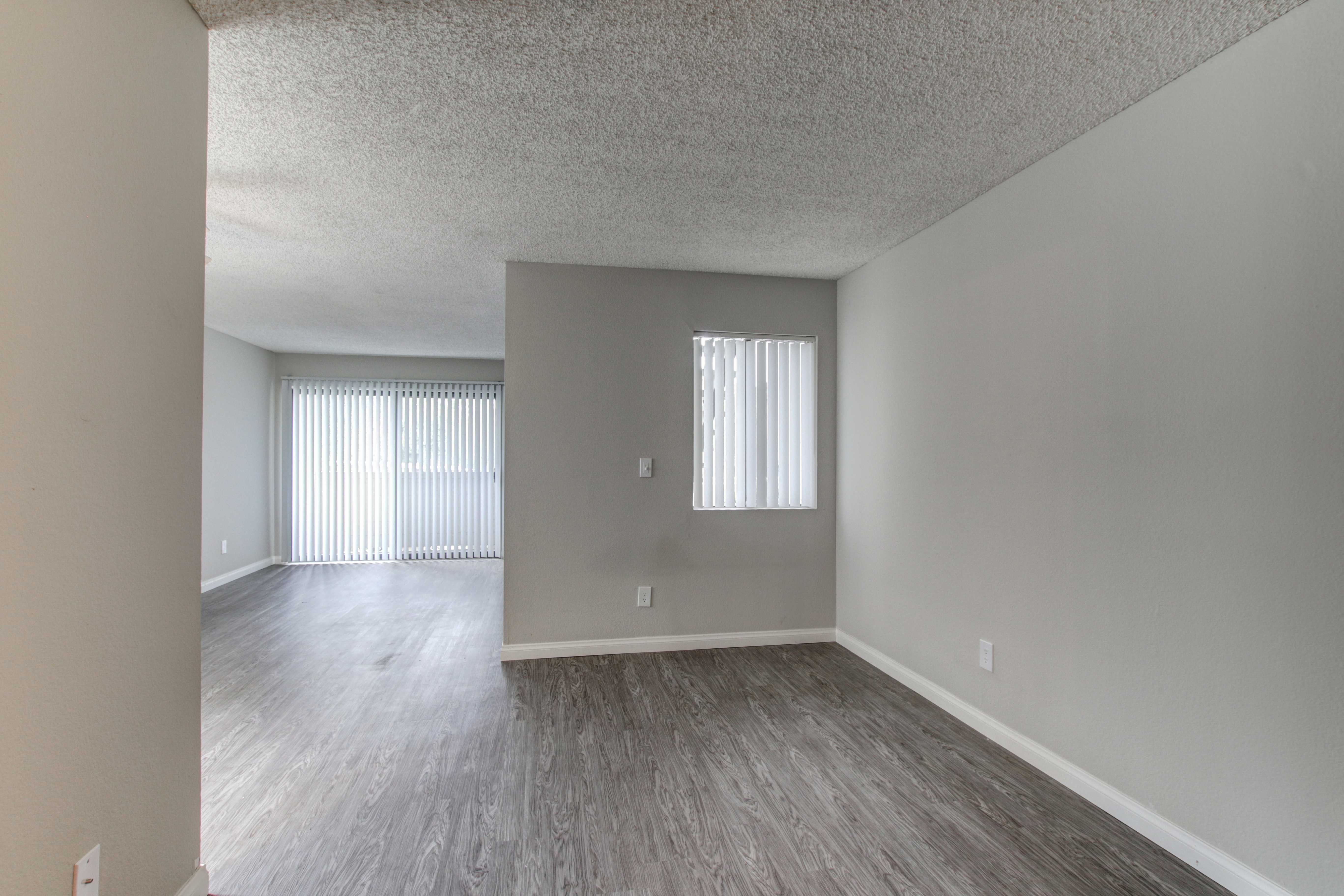 Different angle of unit. This view is from the hallway and adjacent is the living room area. There are two windows, one large and one small, with vertical blinds. There is wooden flooring.
