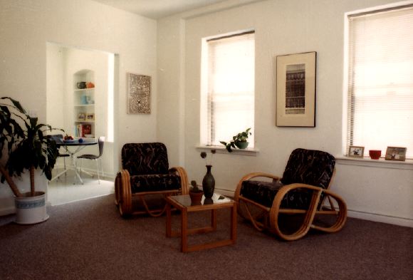 HCHC Recap 1 unit living area. redish carpet in room. two chairs and coffe table. large plant along wall. two large windows