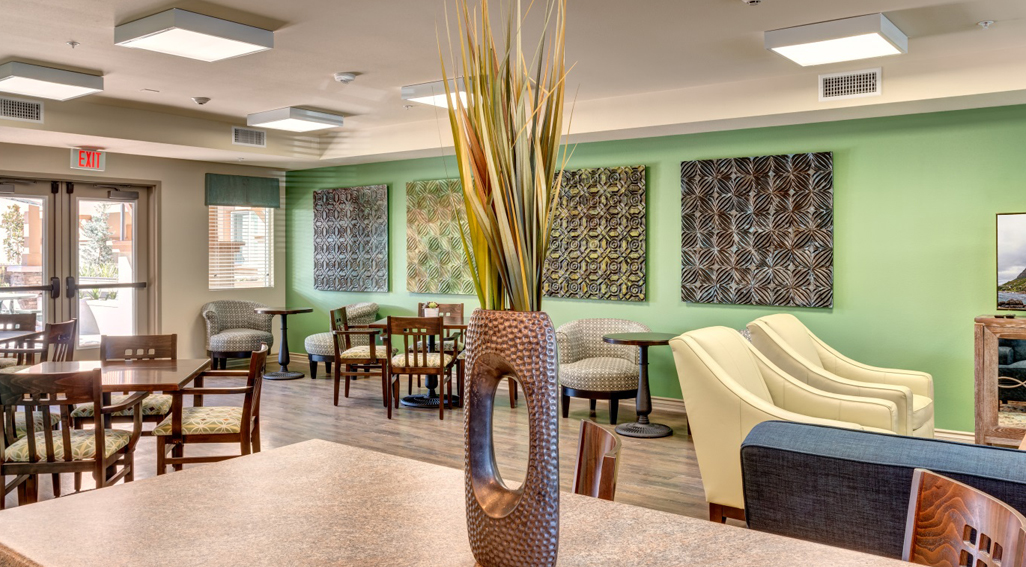 Community room where seniors enjoy events - a dozen wooden chairs, overstuffed chairs and couch, geometric art on walls, plenty of sunlight, large hammered-brass vase with decorative grass