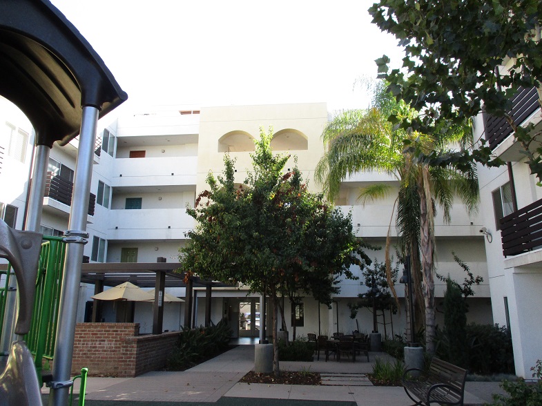 Courtyard of a four story building. There is a bench, multiple seats, plants and trees. Some units have balconies facing the courtyard.
