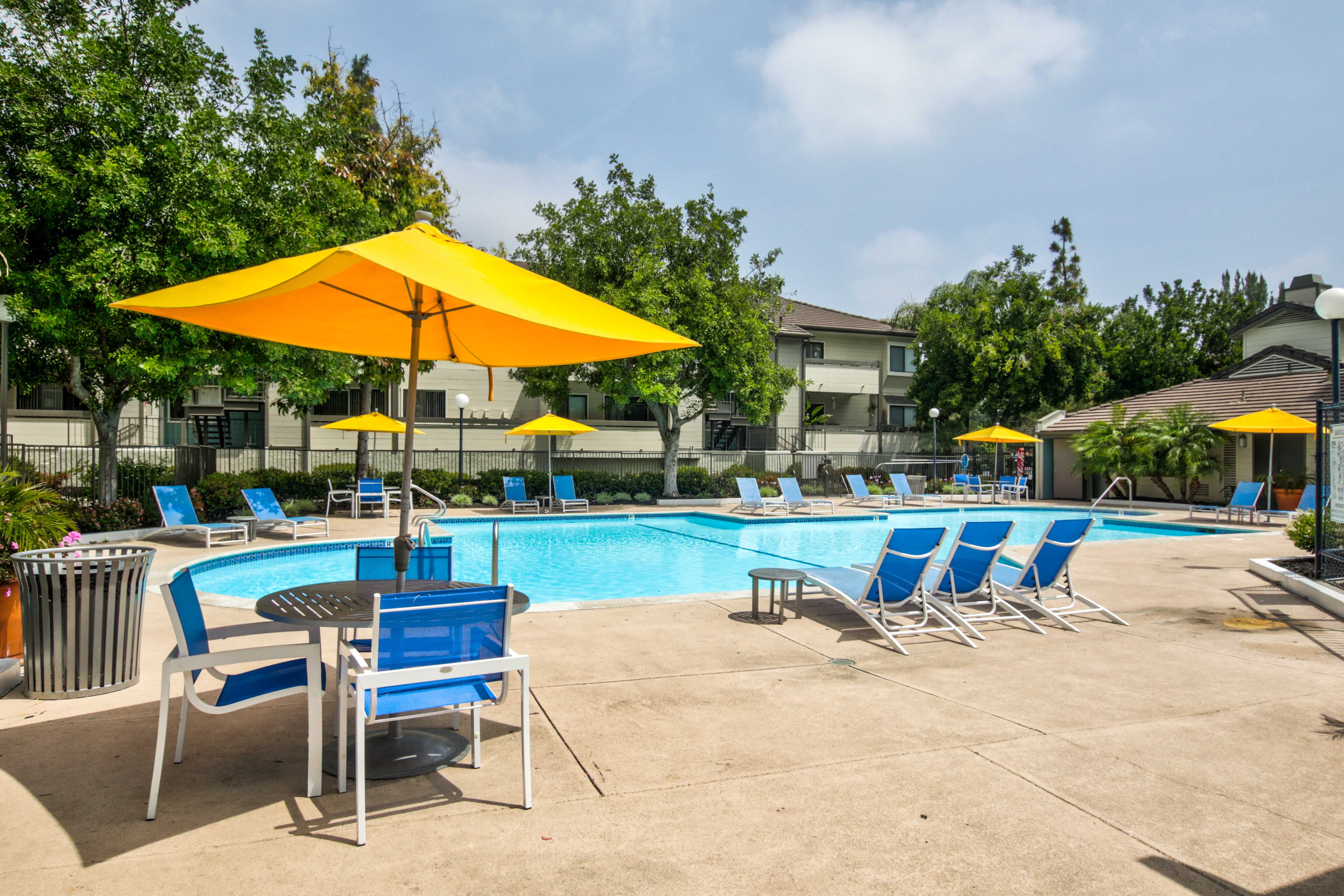 Closer view of pool area that is surrounded by multiple blue lounging chairs, small circular tables, and yellow umbrellas. On the border section of the outter fence is a strip of bushes and trees.