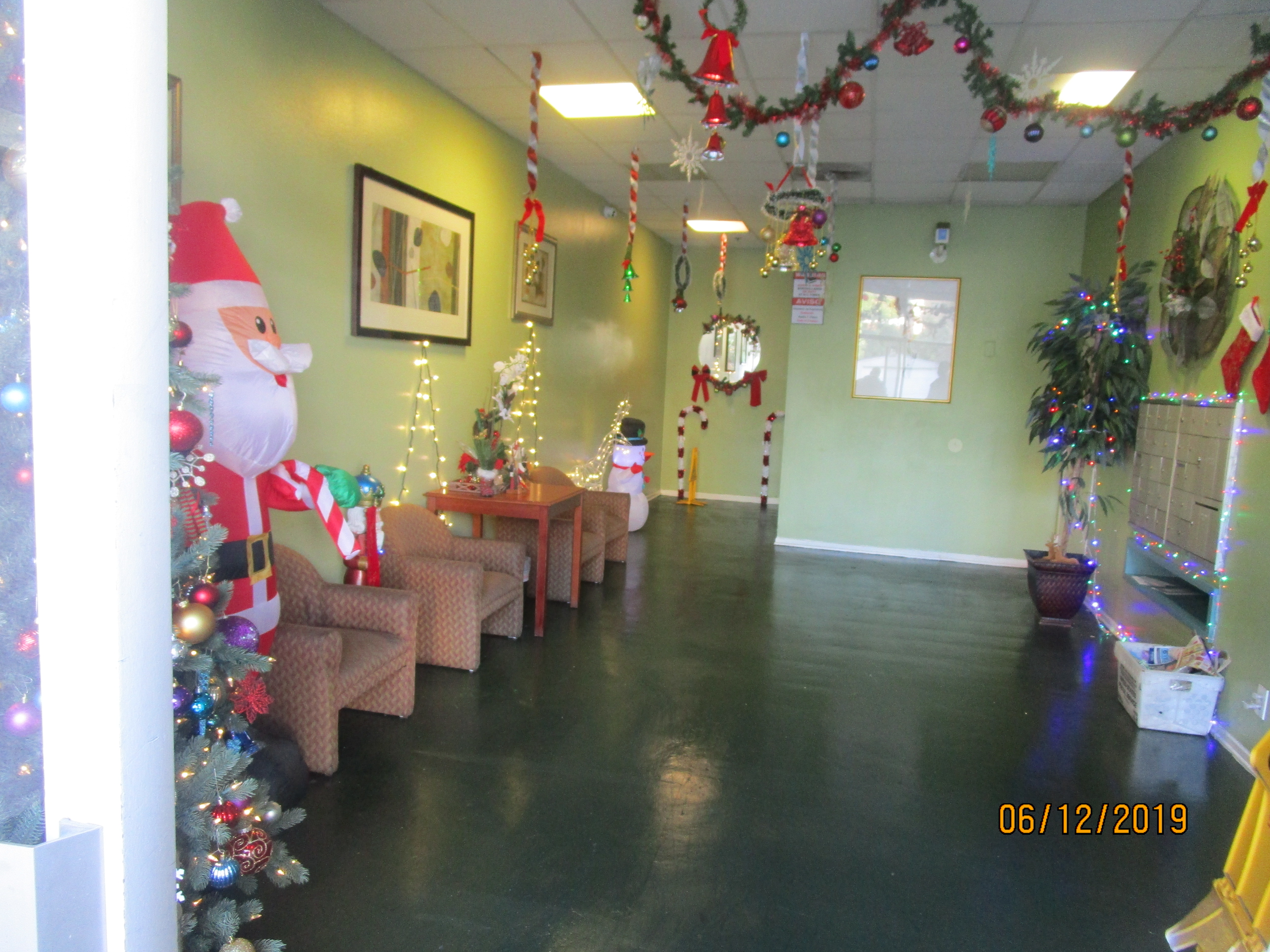 View of a mail room, arm chairs against the light green walls on the left side, a small brown coffee table with Christmas decorations, an inflatable Santa Claus next to a Christmas tree on the left side, an inflatable snowman, tall candy canes and Christm