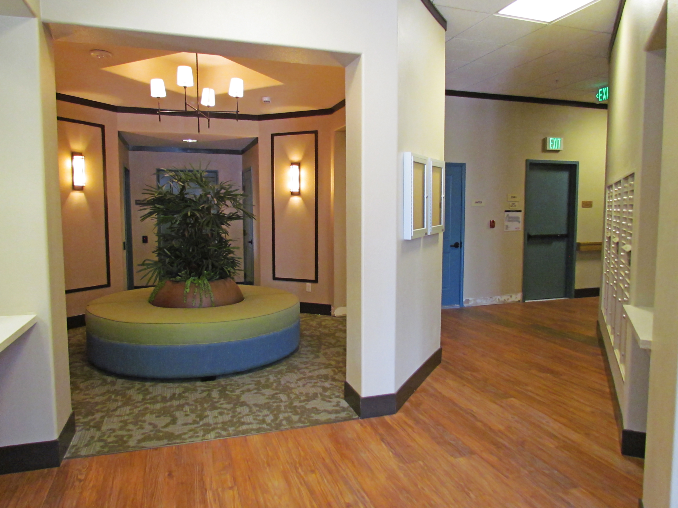 Interior view of the lobby and mail area at Osborne Street Apartments. A circular cushioned seat with a large plant in the center of it in the lobby area and the mail boxes on a wall to the side of the lobby