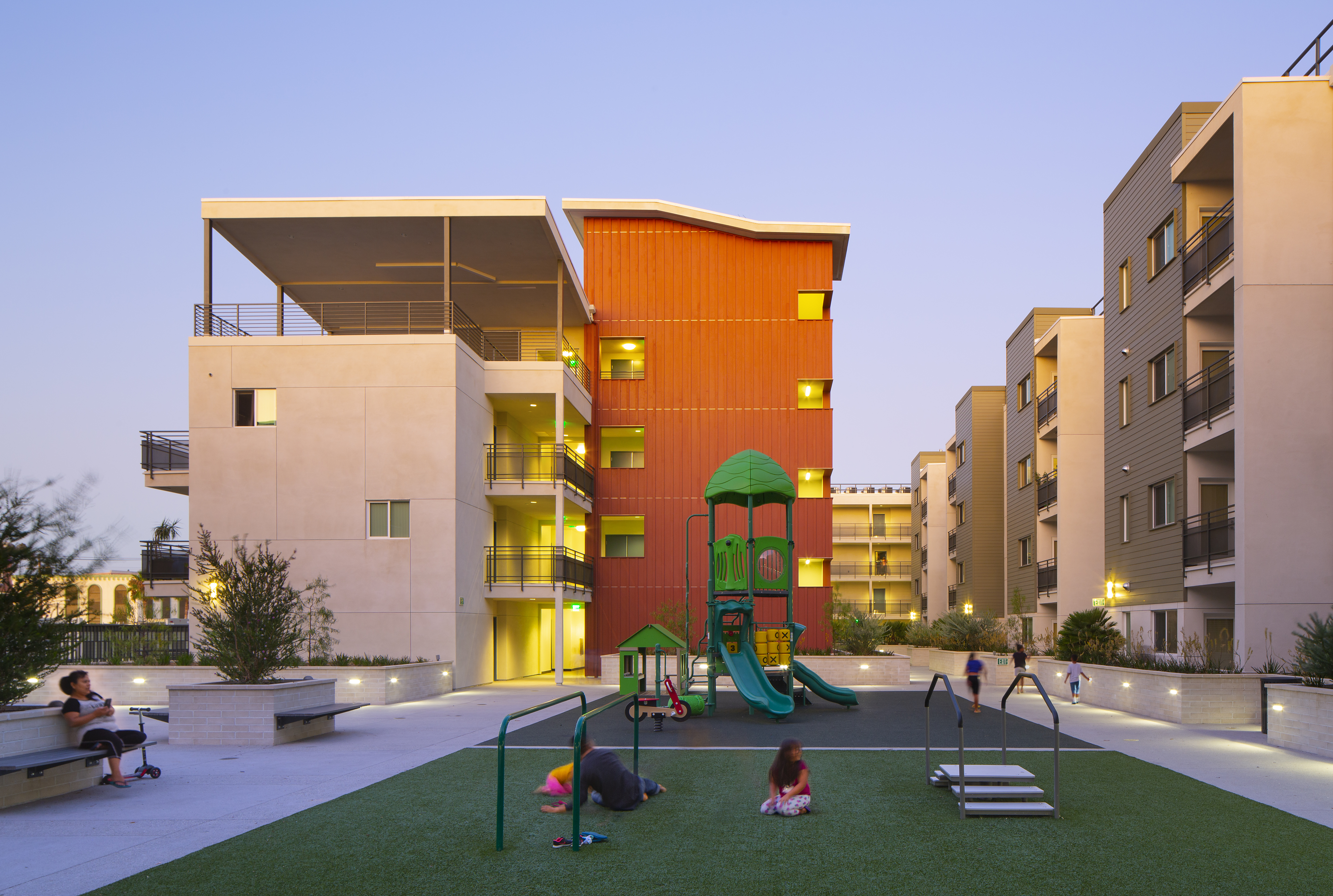 Front view of the playground, view of the building in the back, children playing, a woman with a cell phone on her hand sitting on a bench watching three kids playing, other three kids walking by the playground, gray soft pave flooring on one side and gra