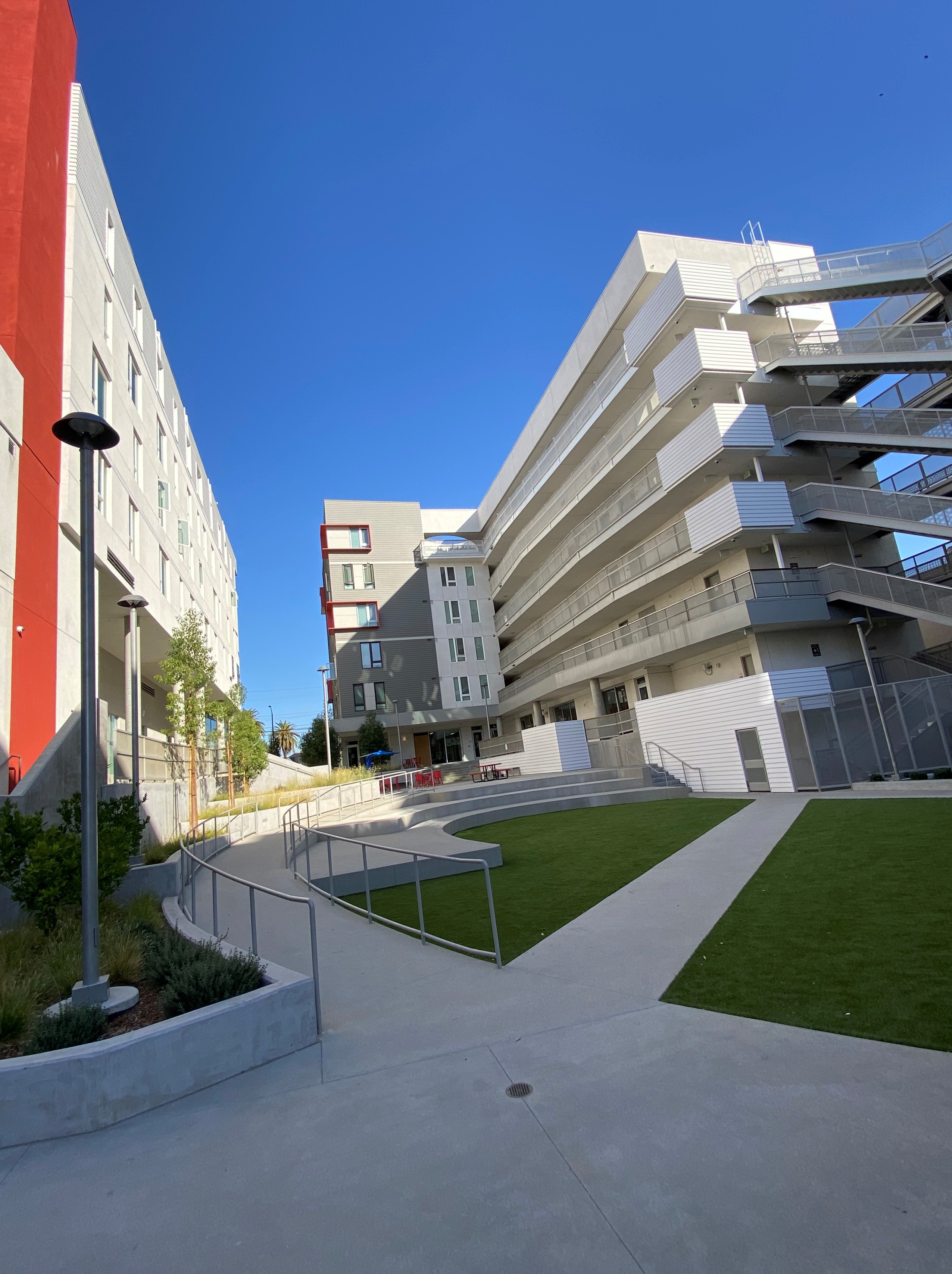 Courtyard with grey multilevel buildings at either side. Grass leading to stairs in the center. A walkway with railing leads to the top of the stairs from the left. Trees in planters line the left building.