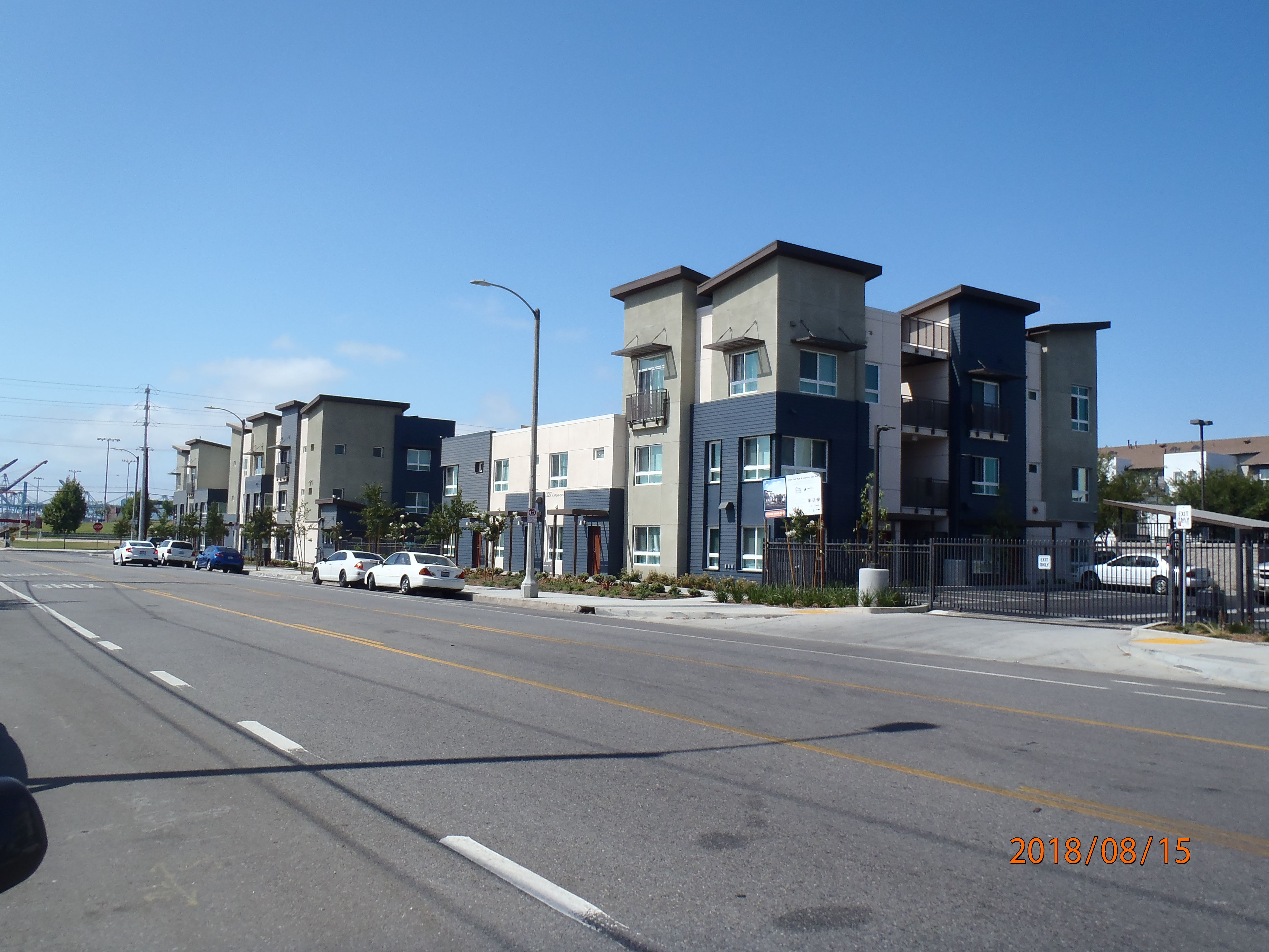 Right side of a multiple and colorful buildings into one, multiple windows, bushes and small palm trees all around, parked cars, a gated open parking visible, wide street view.