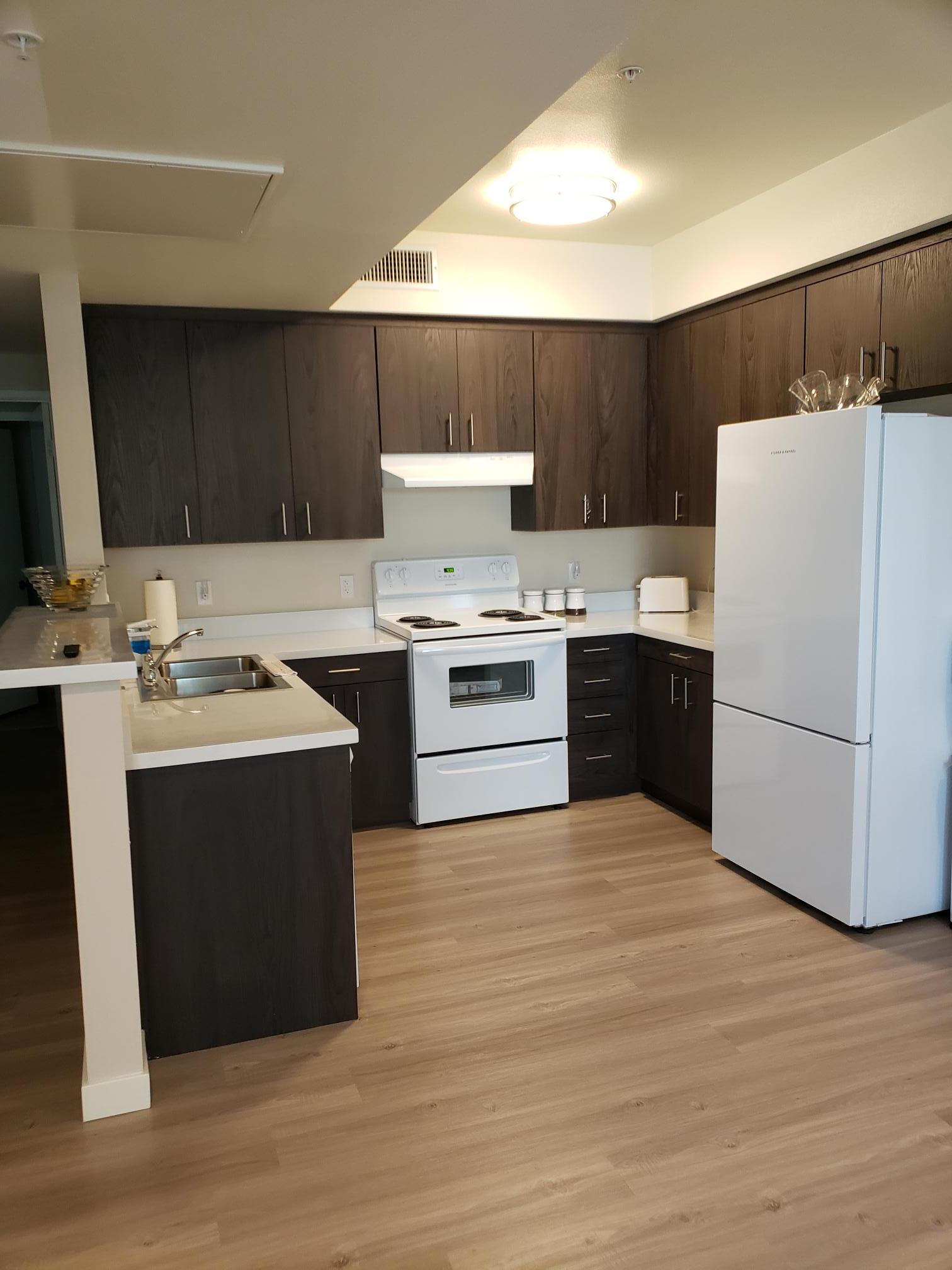 Coronel Apartments unit kitchen. U shape kitchen with dark colored upper and lower cabinets. Full range stove and fridge appliances. Kitchen floors are wood like plants in a light brown tan color.