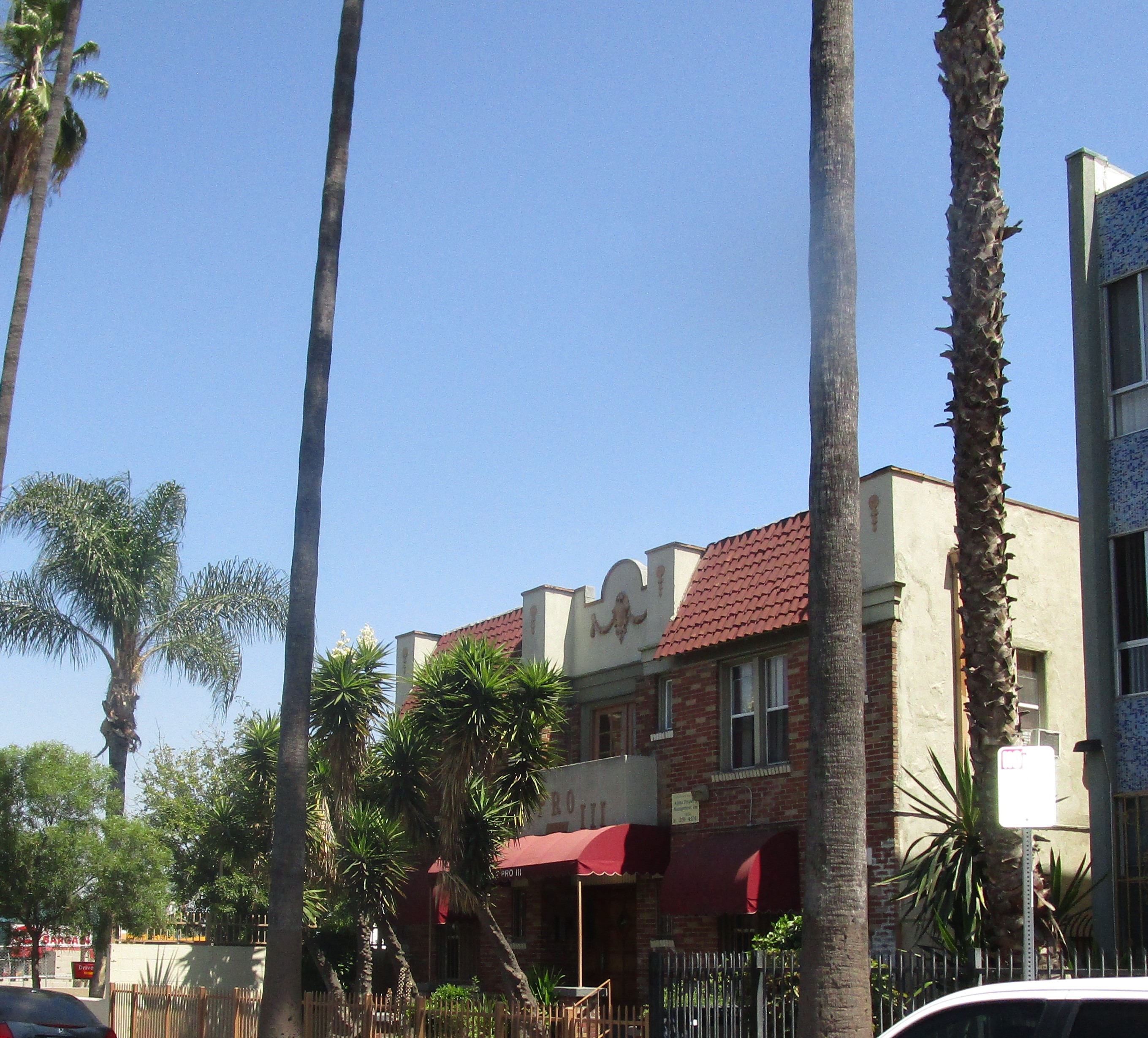 two story beige and brcik color building, one top balcony, main entrance with a burgundy canopy tent, bottom windows on each side with a burgundy canopy tent, Yucca trees right in front of the main entrance, gated, palm trees and other bushes, parked cars