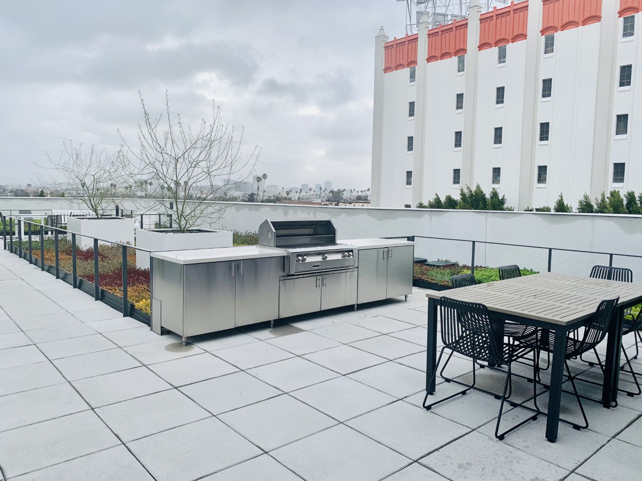 5th floor grill and designated smoking zone. Grill in front of a table with 6 chairs. Trees in planter with flowerbed.