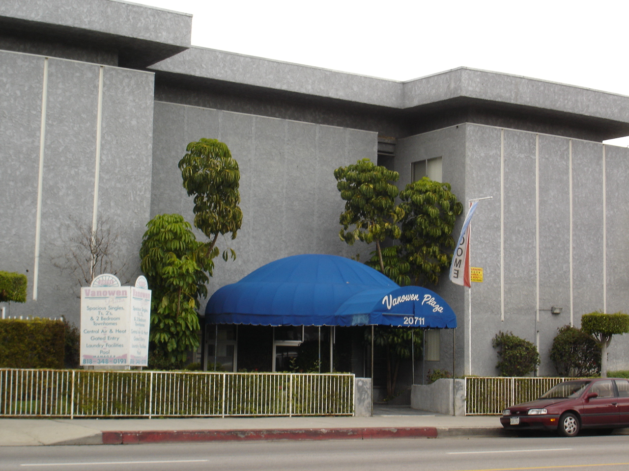 Street view of a gray 2 floor building with a blue cover over the entrance way, trees and green plants decorate outside of the building, red car parked out front