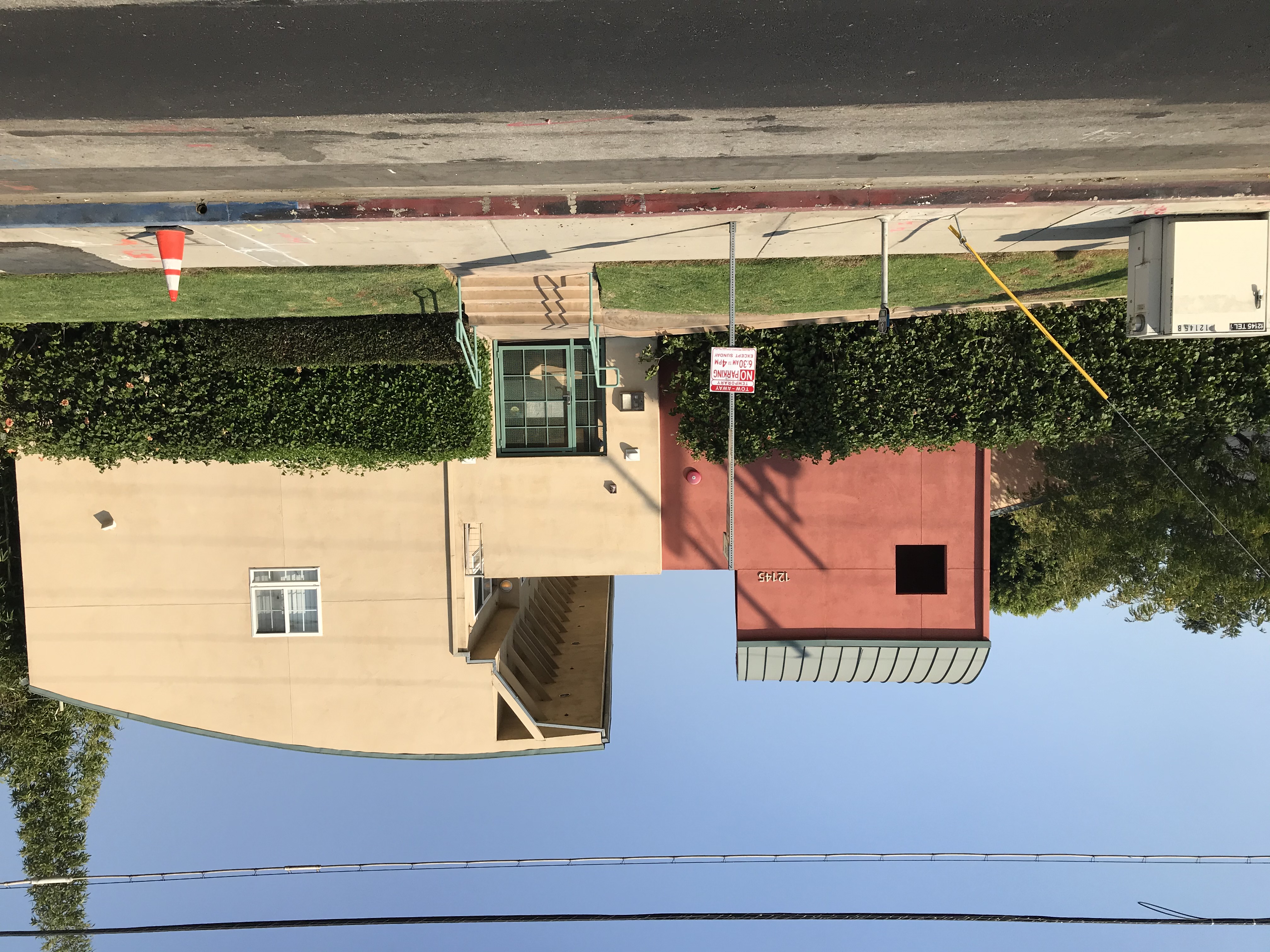 Front view of a three colors two story building, main entrance green iron screen door, four stairs with green handrails, bushes against the walls on each side, temporary no parking sign right in front, disable parking sign, power light big box case, orang