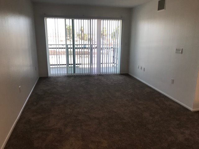 Empty unit consisting of brown carpet, sliding door that leads to a small patio, white vertical blinds, and beige walls.