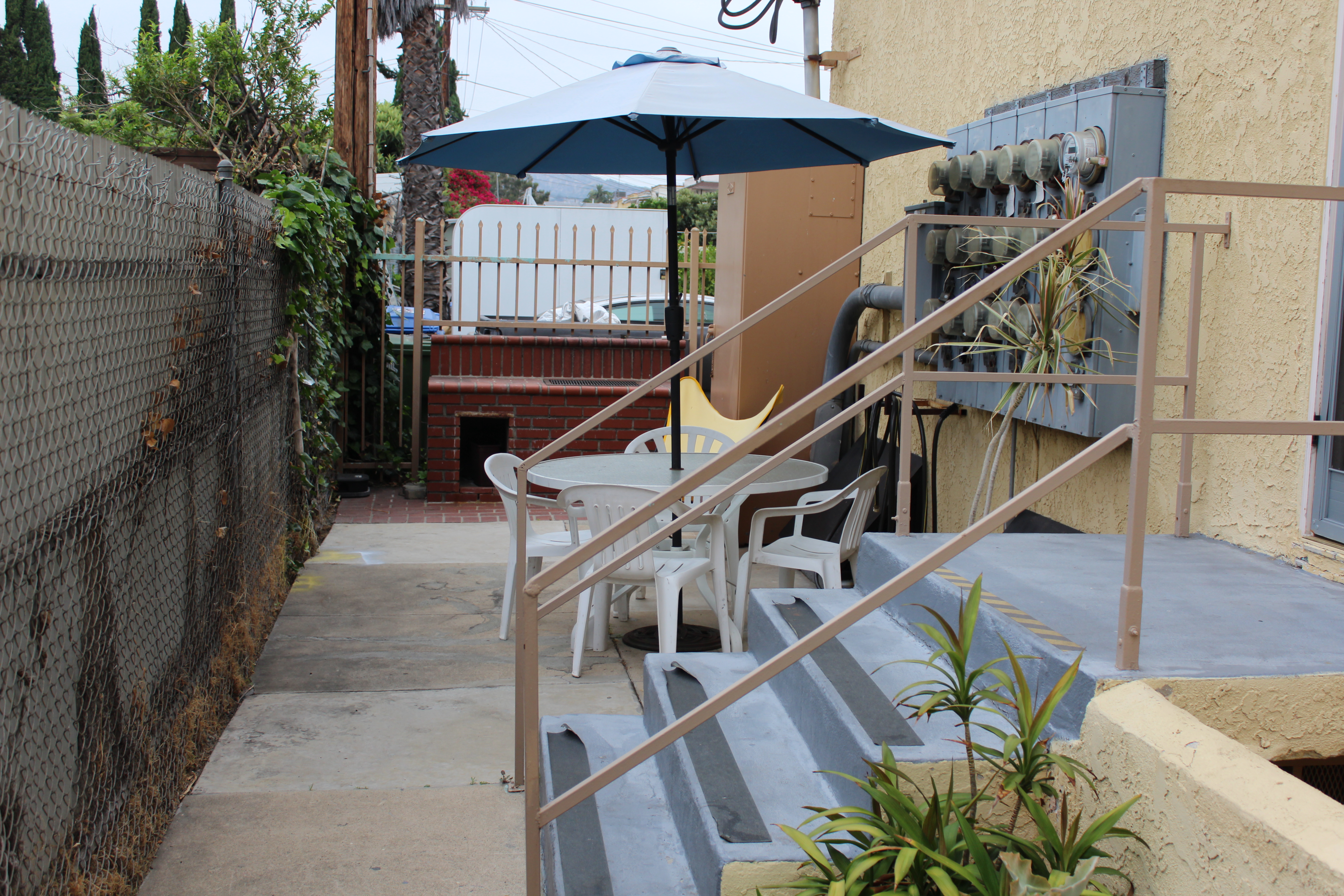 Back patio with a table. chairs, and outdoor umbrella next to electrical panel and stairs with hand rails.
