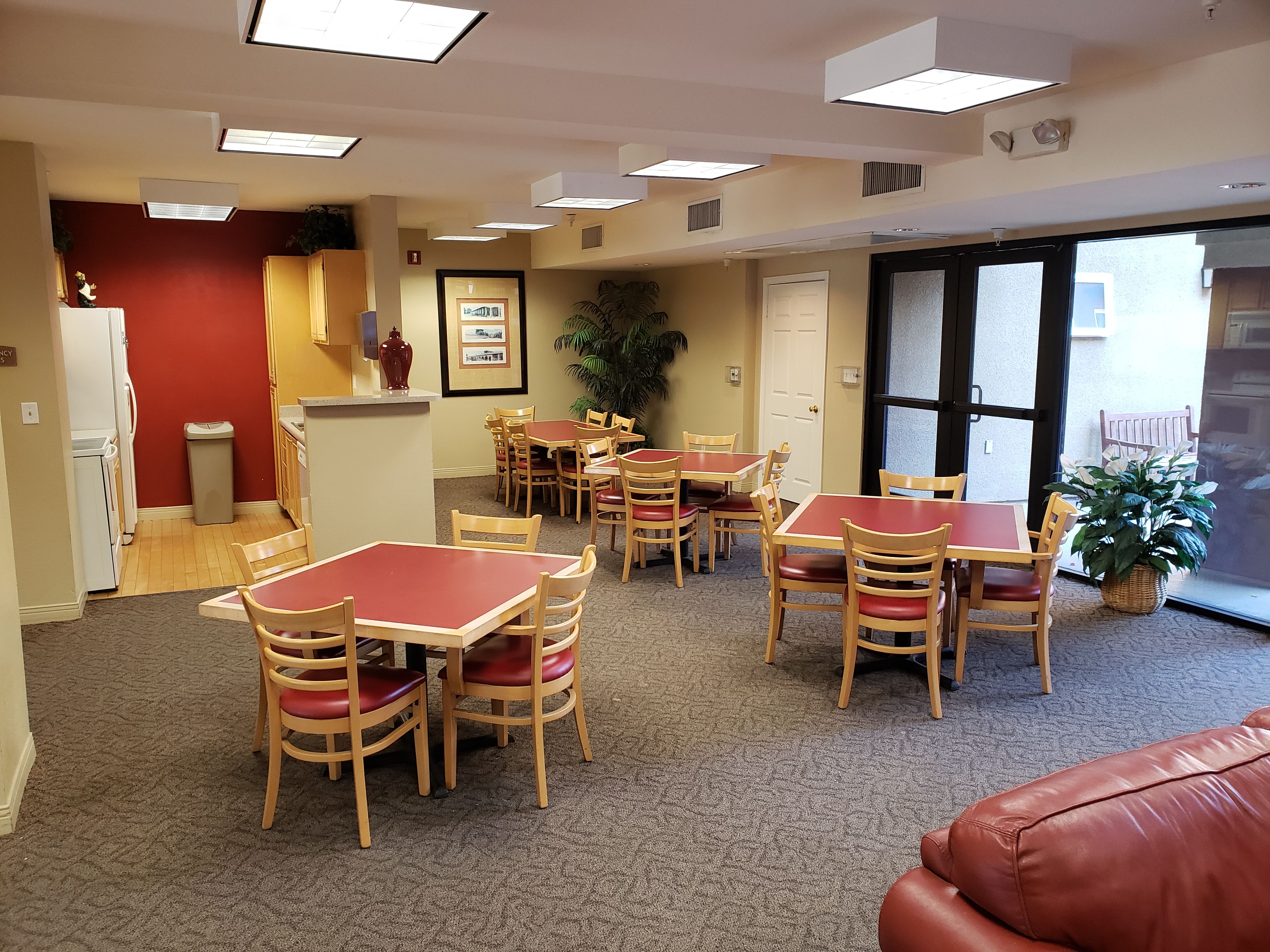 Image of vintage crossing senior apartments community room. Lots of lighting across ceiling. Large room with kitchen and several tables and chairs for lounging. Large glass double doors and windows that bring in a lot of natural lighting. Kitchen applianc