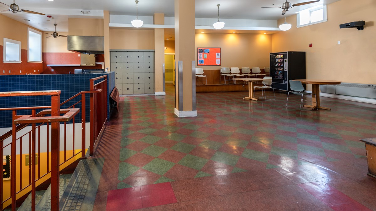 View of a room, burgundy and green tile floor, stairs with handrailson the left side, two round tables with one chair each, six beige chairs against the wall toward the end of the room, big black vending machine, orange square board on the wall, white lig