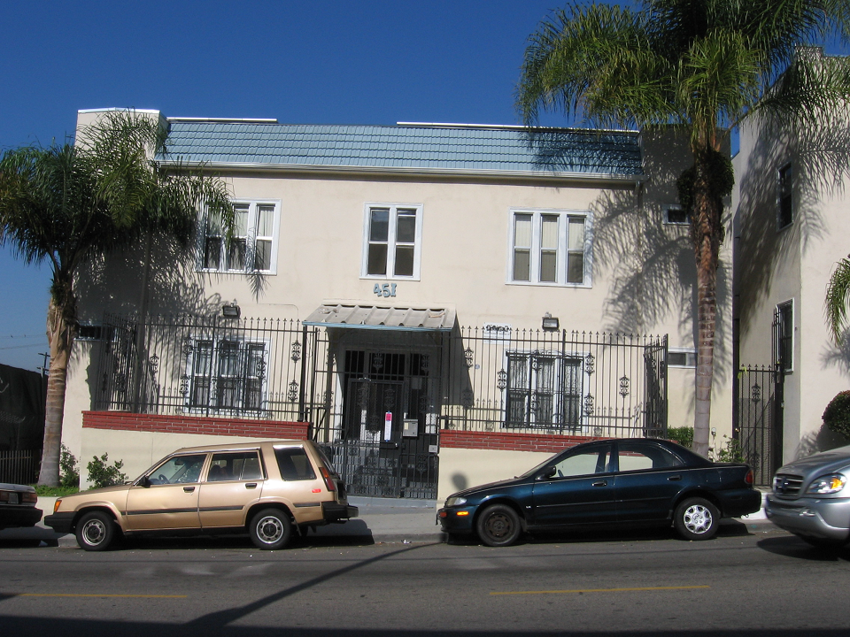 Front view of a beige two story building, multiple windows with white trim, gated building, bottom windows with security bars, gray steps toward the main entrance, call box on the right side, parked cars in front, palm trees and plants.