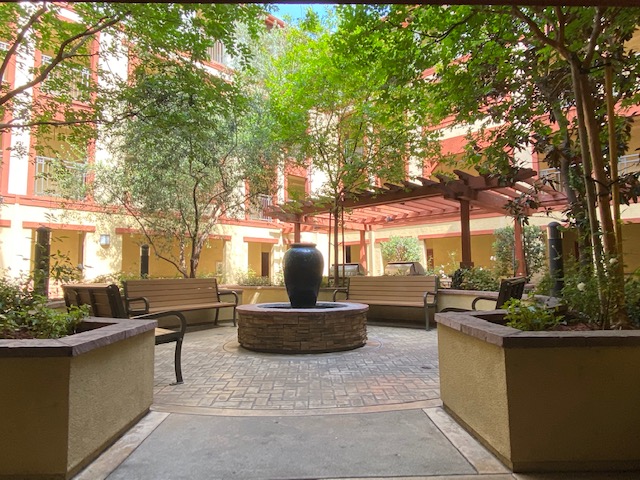 Exterior view of a courtyard at Sunrise Apartments. Circular area with fixed wooden benches around a fountain, and large garden boxes surrounding the benches