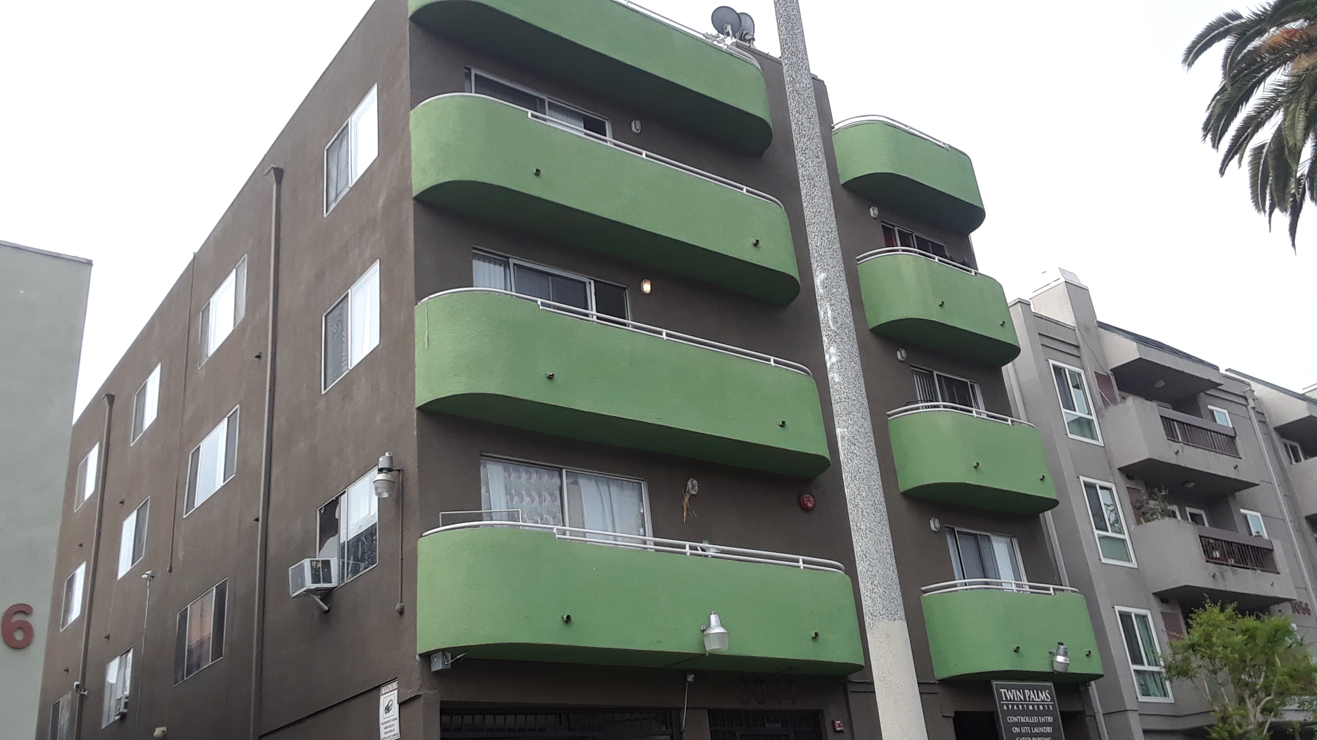 Front angle view of a four story building. Units facing the street have green balconies. The fronds of a very tall date palm on the sidewalk are just visible.