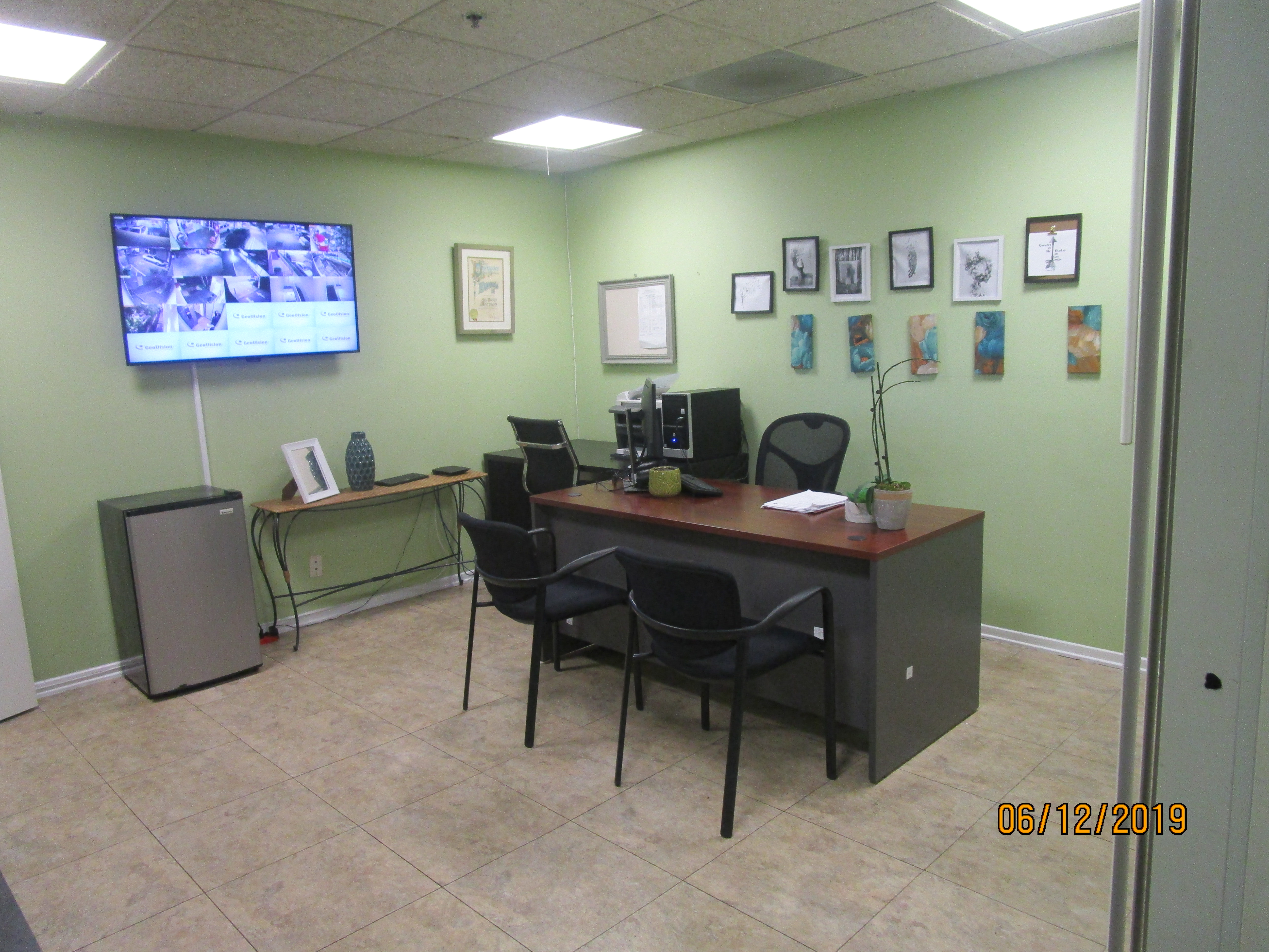 View of an office, light brown and beige linoleum floors, light green walls, multiple picture frames, a TV hanging on the left side wall, a compact fridge, a coffee table with a picture frame and decorations against the left side wall, a desk with a chair