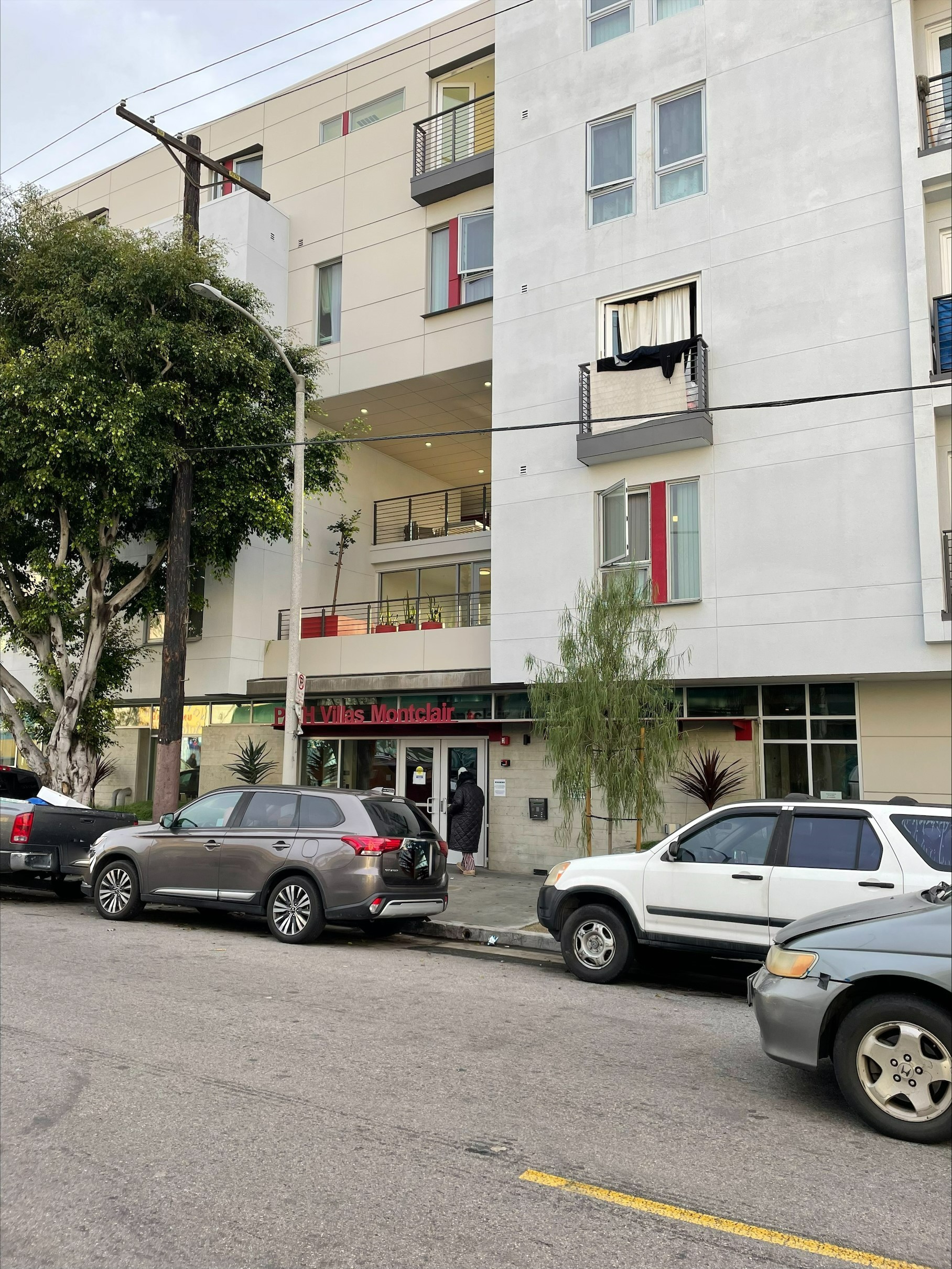 Street view of building. Cars parked in front of building. Balconies can be seen above the buildings entrance. Two trees on the sidewalk.